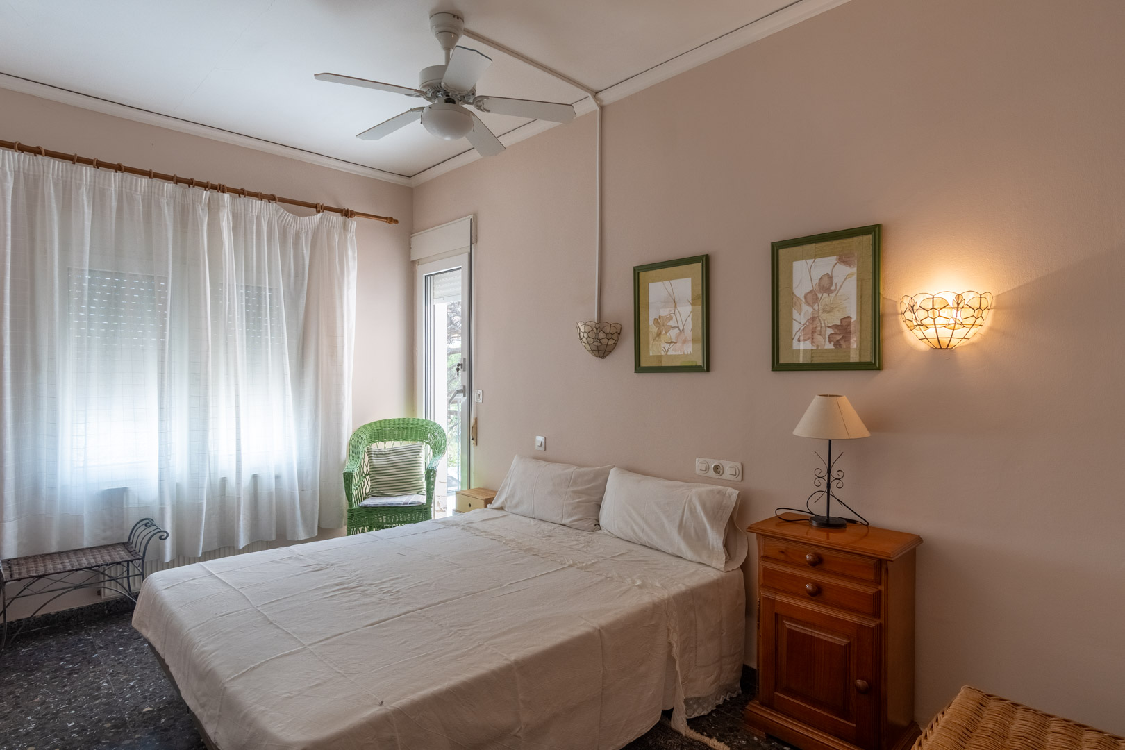 Bedroom featuring a double bed, ceiling fan, and direct outdoor access. Includes wooden furniture, terrazzo flooring, and a decorative wicker chair by the window.