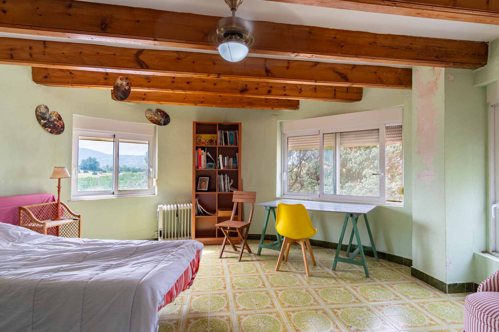 Bright bedroom featuring exposed wooden beams, patterned ceramic floor tiles, and a dedicated workspace. Large windows offer open views of the surrounding Mediterranean landscape.