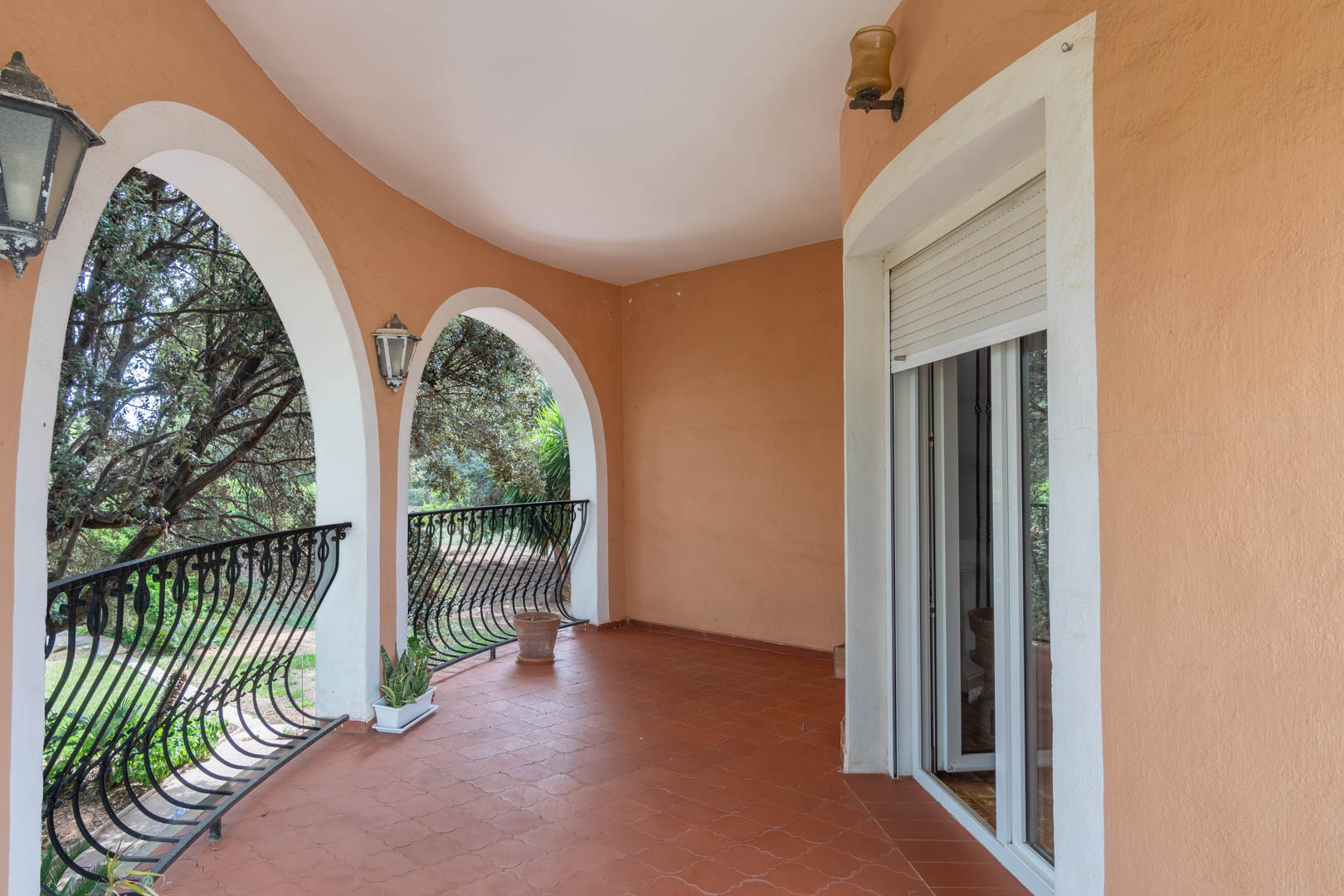 Covered terrace featuring Mediterranean arches, terracotta tile flooring, and wrought-iron railings overlooking the lush garden.