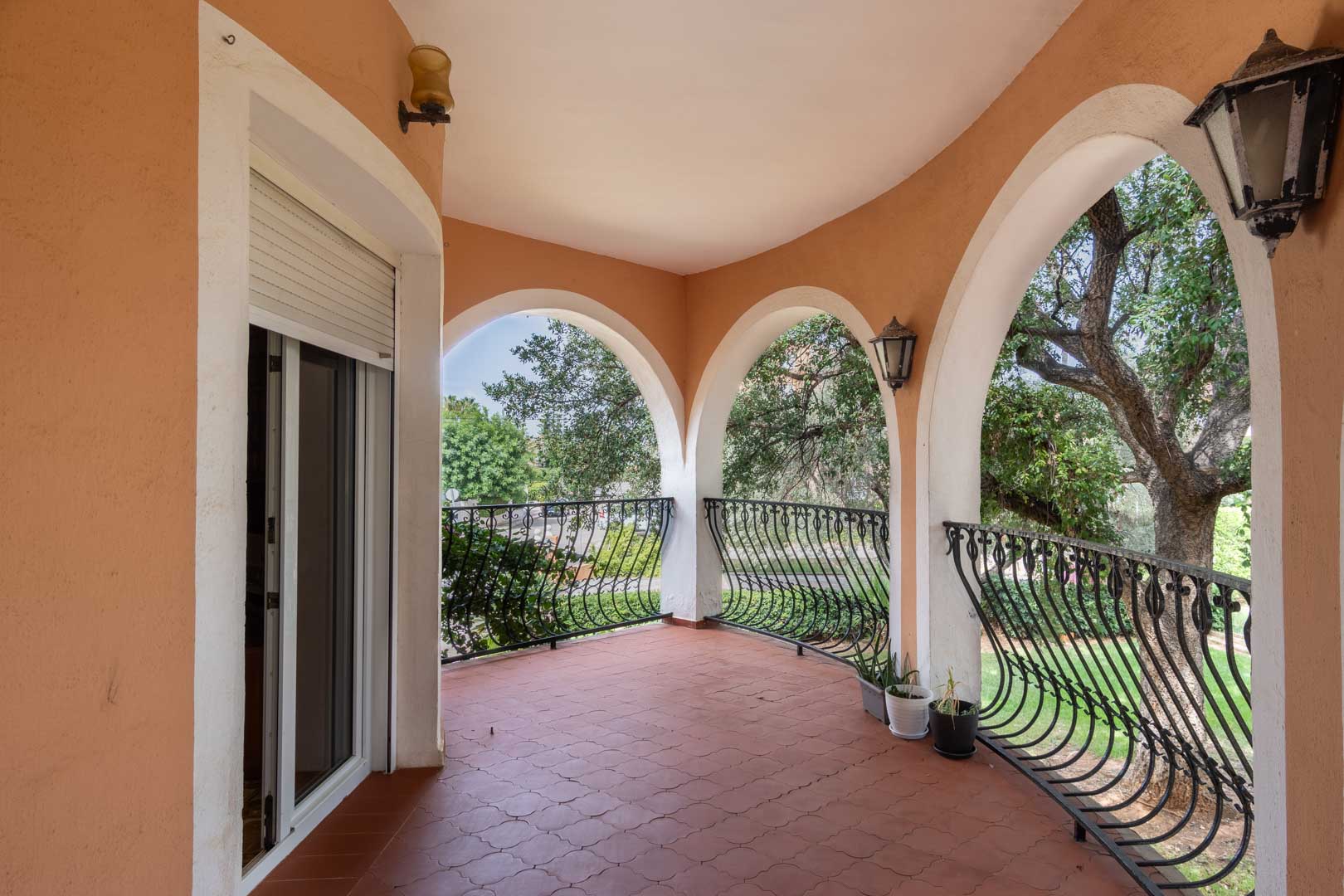 Covered terrace featuring white Mediterranean arches, curved wrought-iron railings, and terracotta tile flooring overlooking the mature garden.