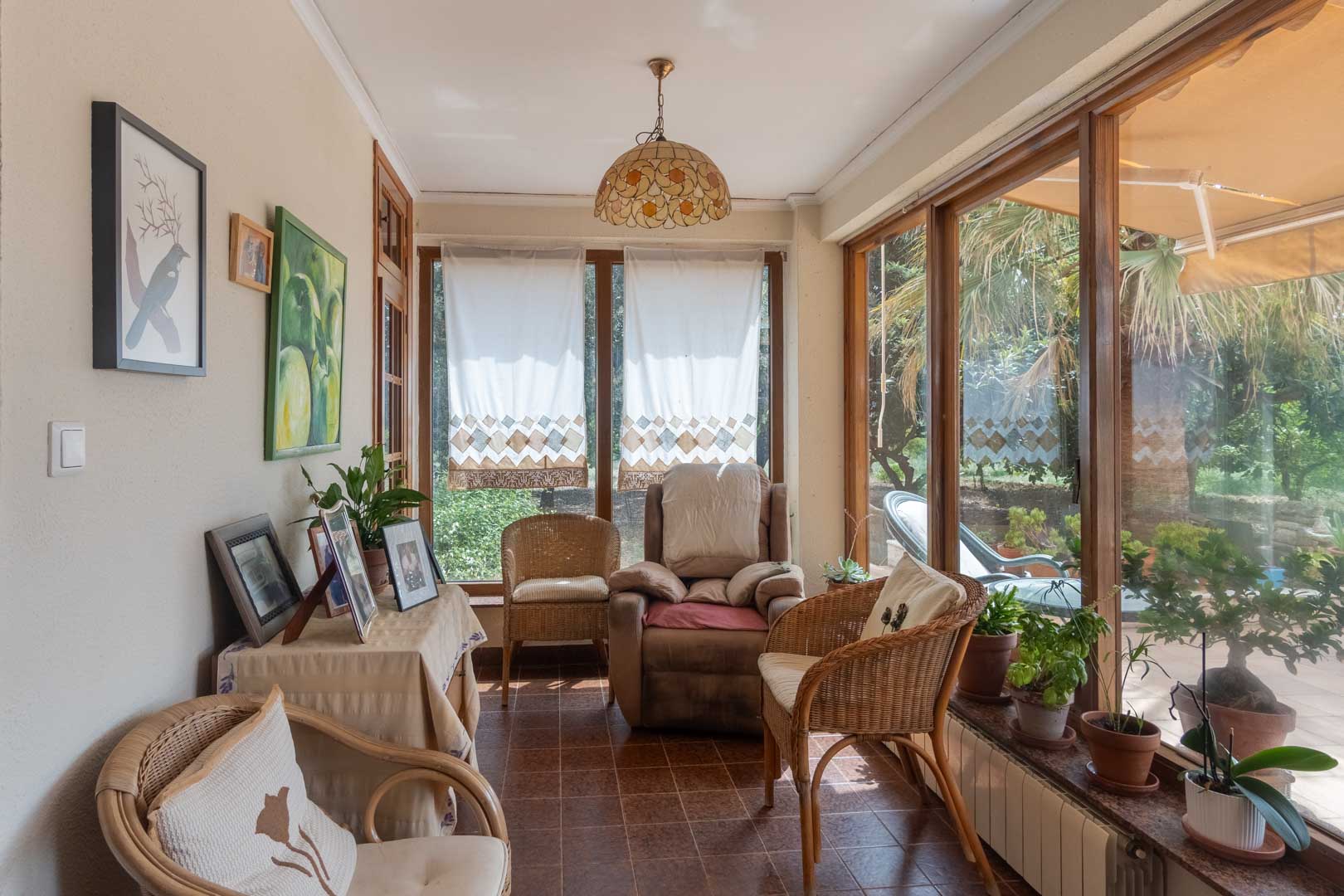 Enclosed sunroom with wooden-framed windows and ceramic tile flooring. This bright space offers garden views and a comfortable wicker seating area.