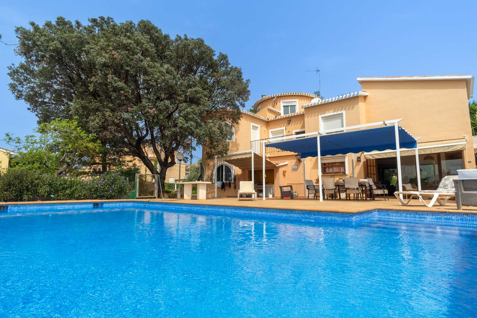 Exterior view of the private swimming pool and sun terrace, featuring the Mediterranean-style villa with a shaded outdoor dining area under a blue awning.