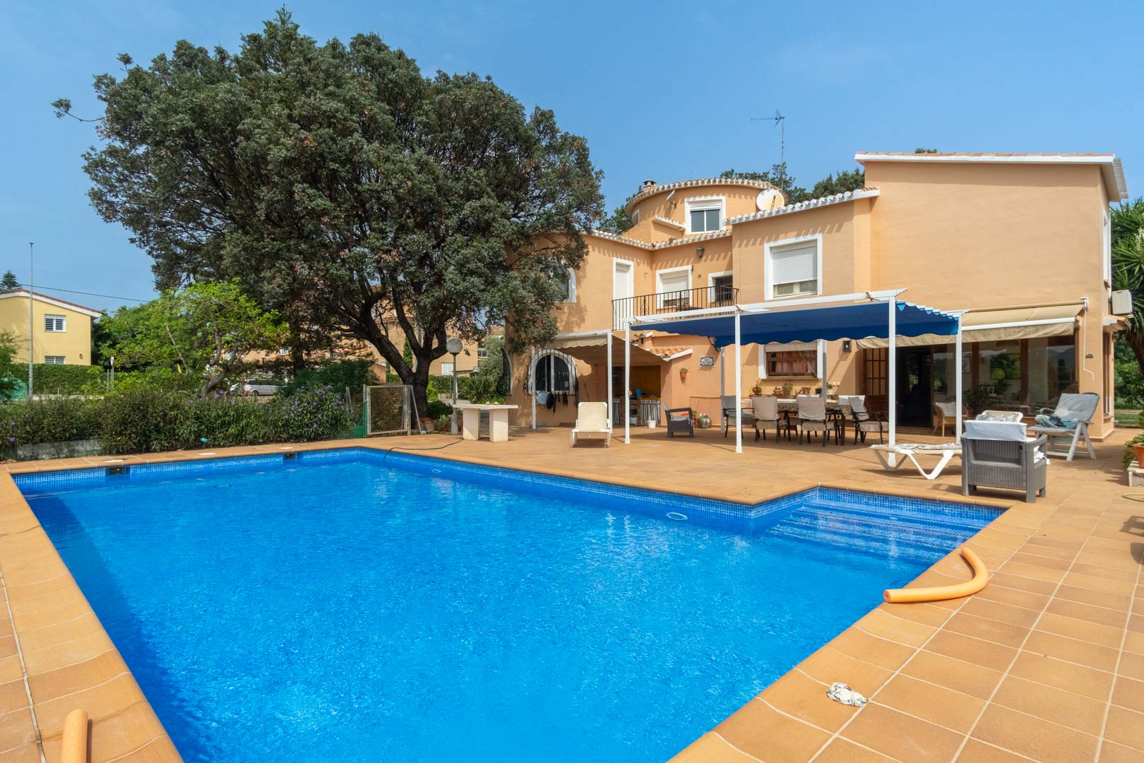 Exterior view of the rectangular swimming pool with a terracotta tile surround, featuring the Mediterranean villa facade and a shaded dining area with a blue awning.