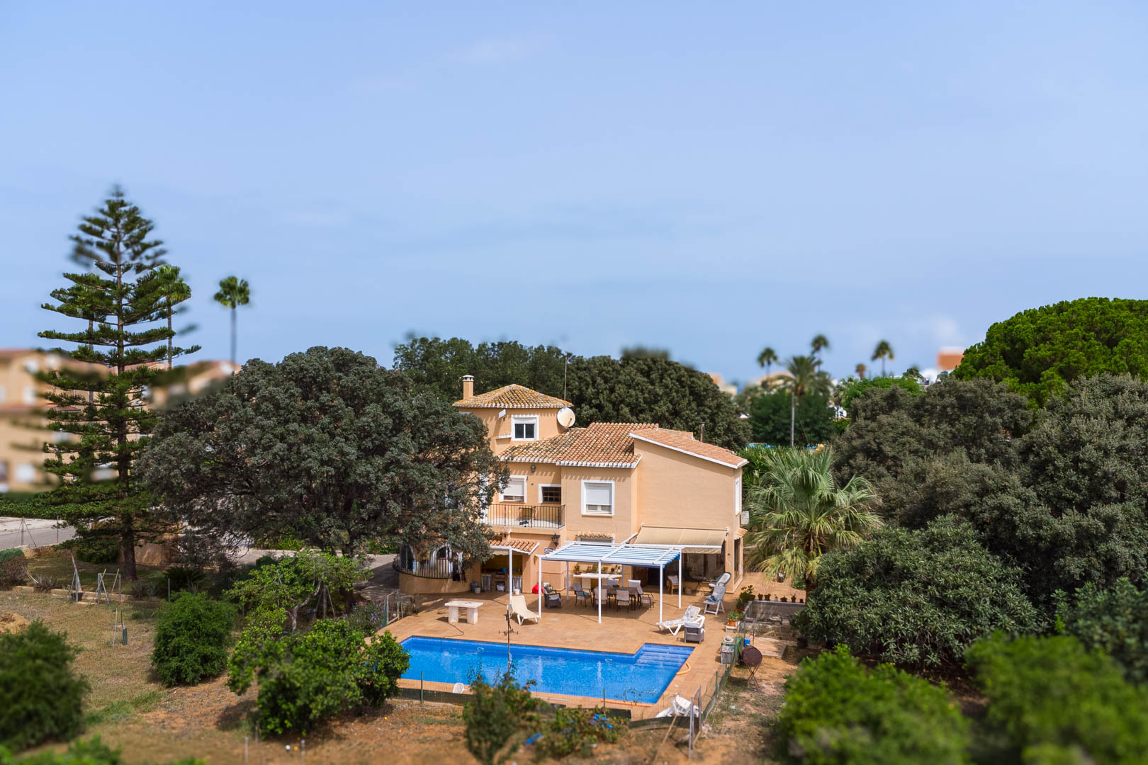 Exterior view of the villa featuring a rectangular swimming pool, a white pergola-shaded terrace, and outdoor dining areas set within a lush Mediterranean garden.