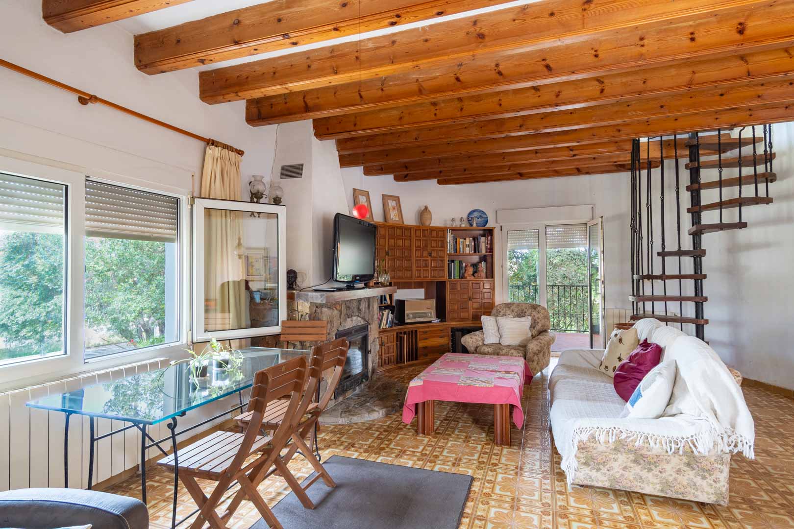 Living area featuring exposed wooden ceiling beams, a stone fireplace, and a spiral staircase leading to the upper level, complemented by tiled floors.