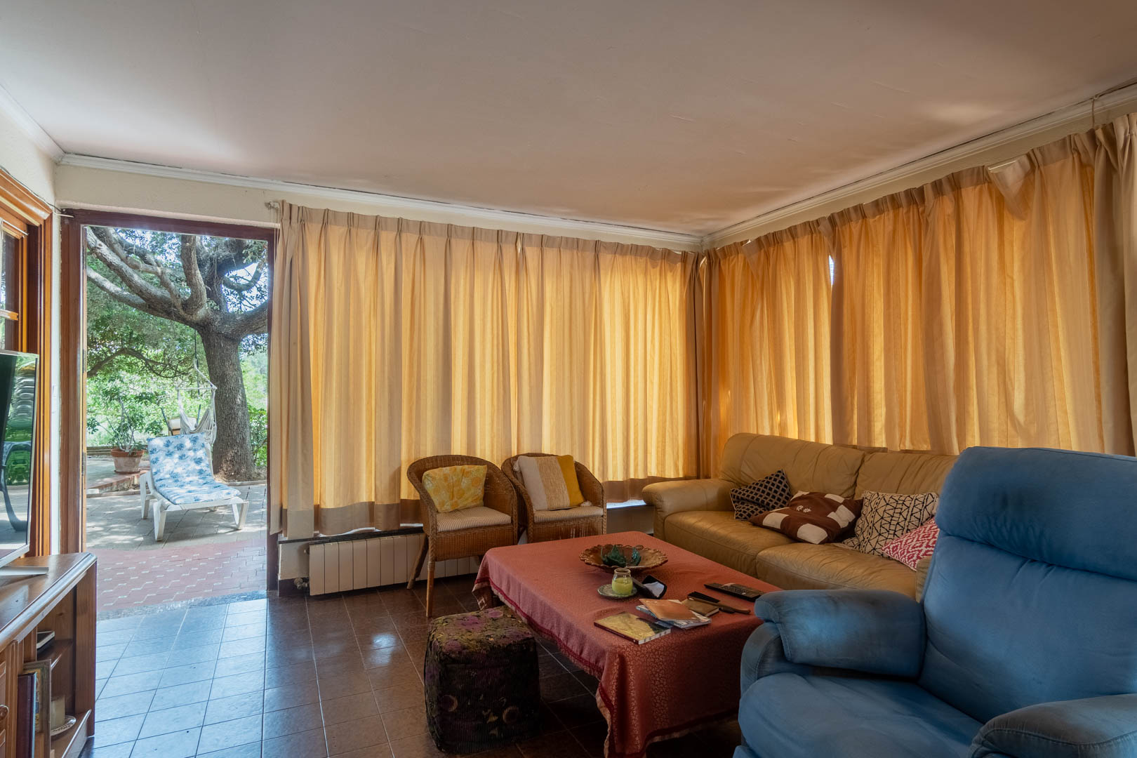 Living room with ceramic tile flooring and direct access to a paved terrace featuring a mature tree. Large curtains and a radiator complement the functional layout.