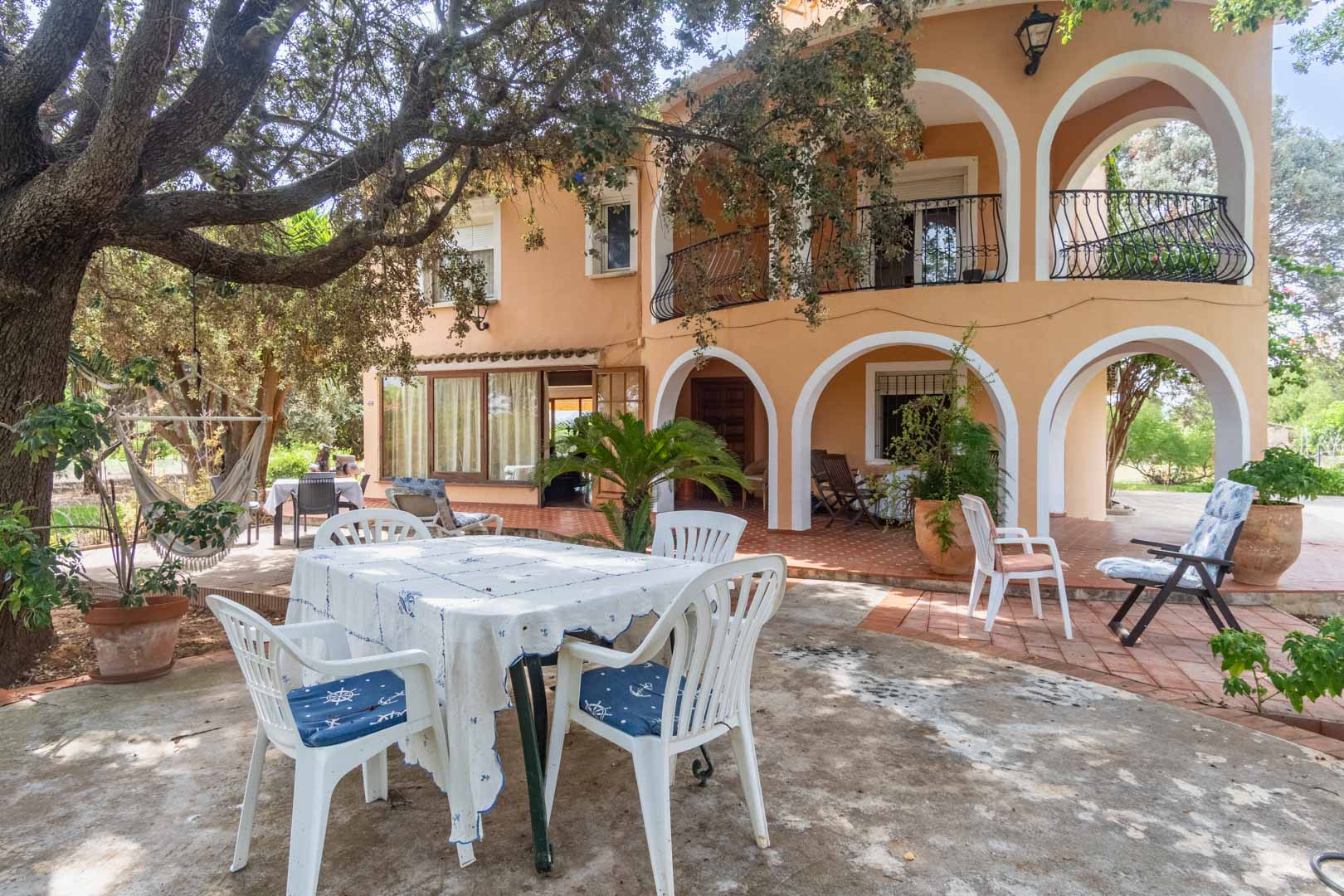 Mediterranean facade featuring white arched porches, spacious terraces shaded by mature trees, and outdoor dining areas on terracotta flooring.