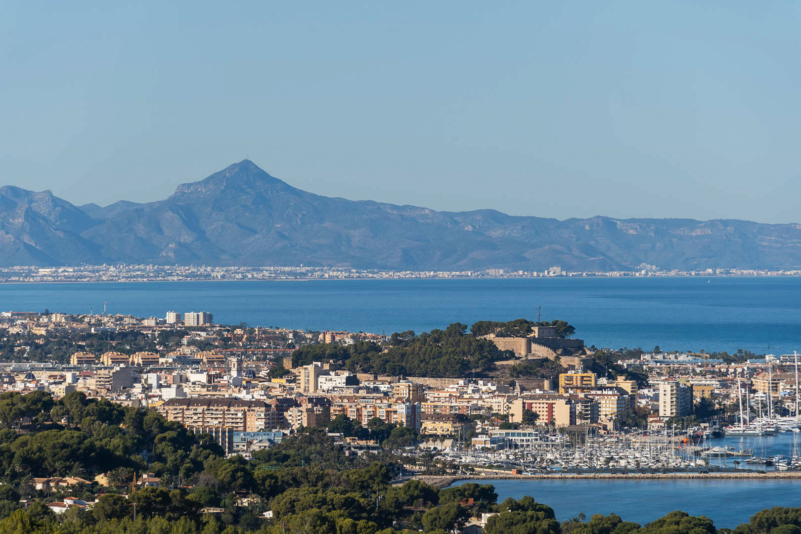 Panoramic view of Dénia featuring the historic castle, the marina filled with yachts, and the Mediterranean coastline set against a backdrop of distant mountains.