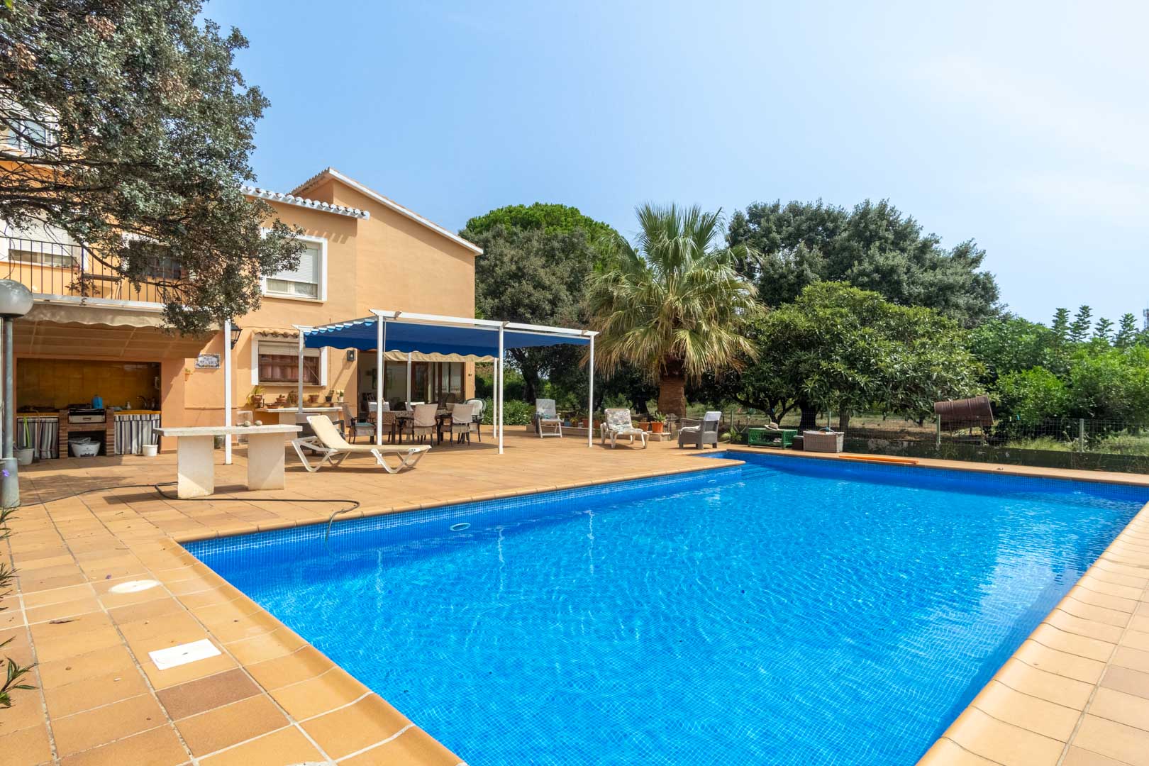 Rectangular swimming pool with blue mosaic tiling, featuring a spacious terracotta terrace, outdoor kitchen, and a shaded dining area with a blue awning.
