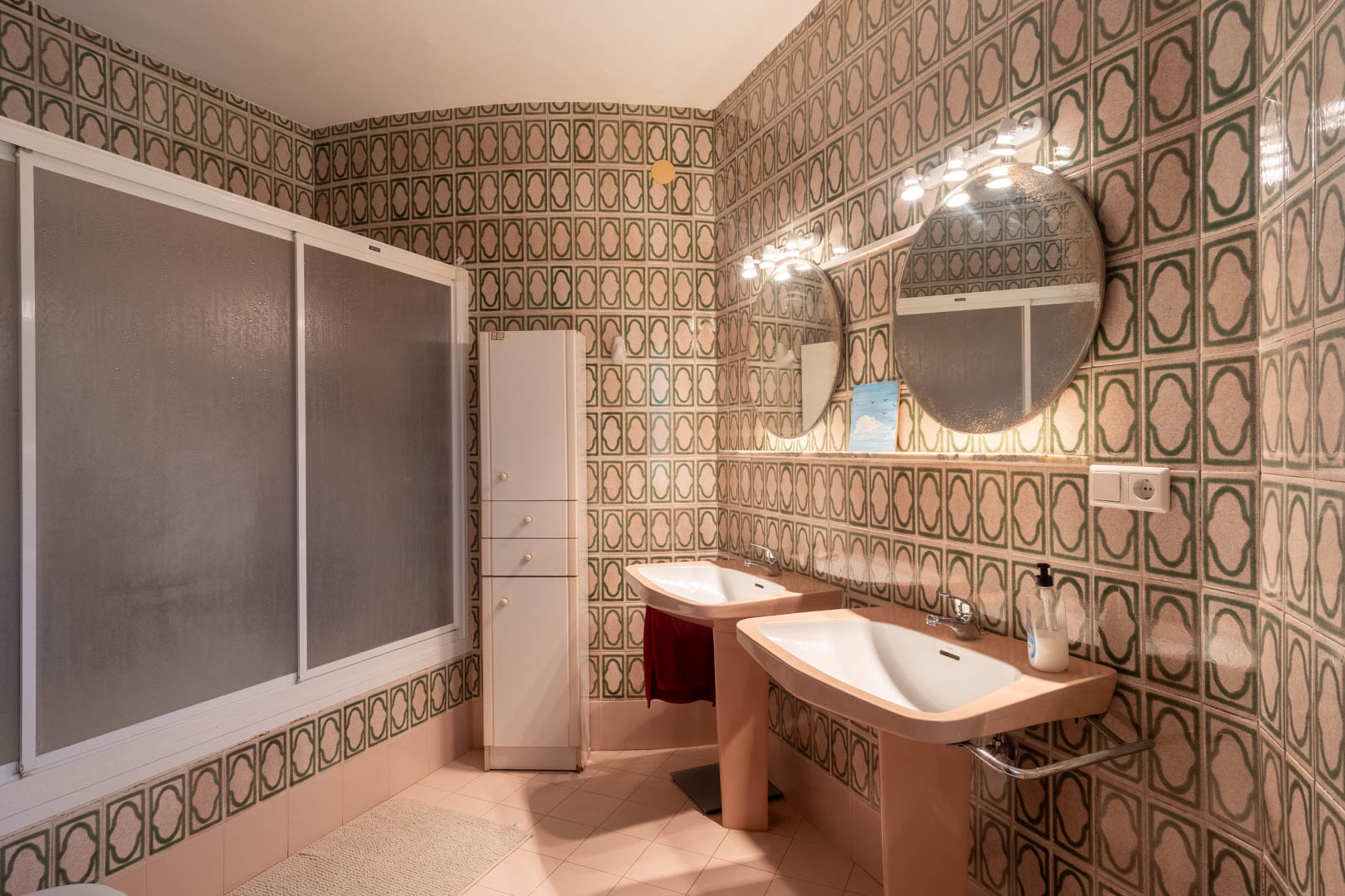 Retro-style bathroom featuring double pedestal sinks, illuminated circular mirrors, and green patterned wall tiles. Includes a walk-in shower with frosted sliding doors.