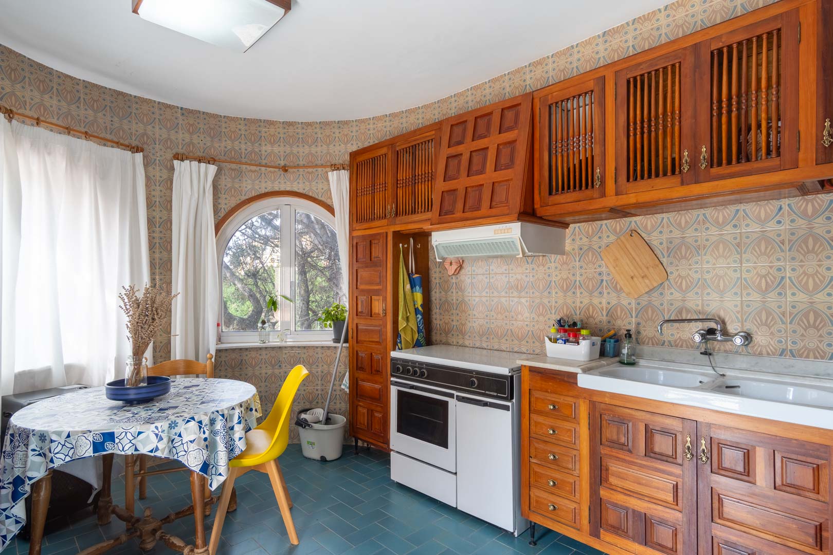 Rustic kitchen featuring carved wooden cabinetry, decorative patterned wall tiles, and a breakfast nook by an arched window overlooking the garden.