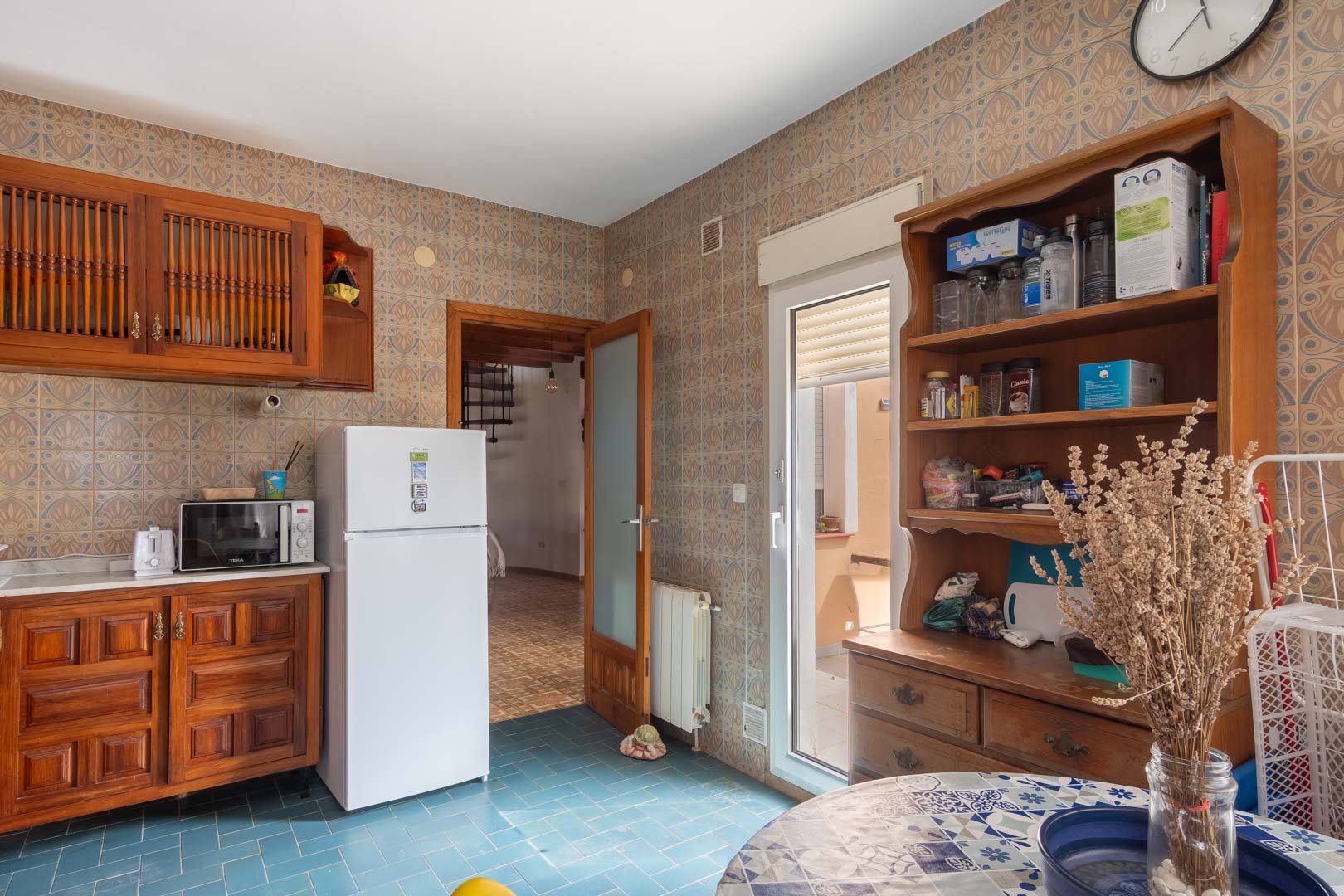 Rustic kitchen featuring wooden cabinetry, blue tiled floors, and patterned wall tiles. The space includes a glass door to the exterior and views of the adjacent living area.