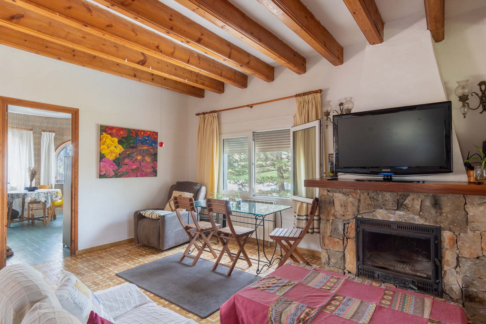 Rustic living area featuring exposed wooden ceiling beams, a natural stone fireplace, and a glass dining table set against tiled flooring.
