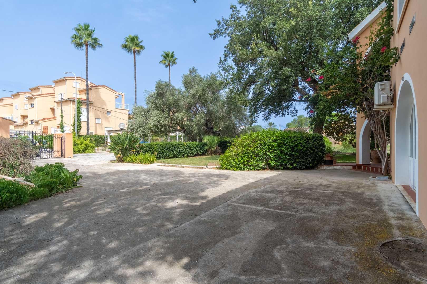 Spacious paved driveway and entrance area featuring wrought iron gates, mature Mediterranean trees, palms, and traditional arched architectural details.