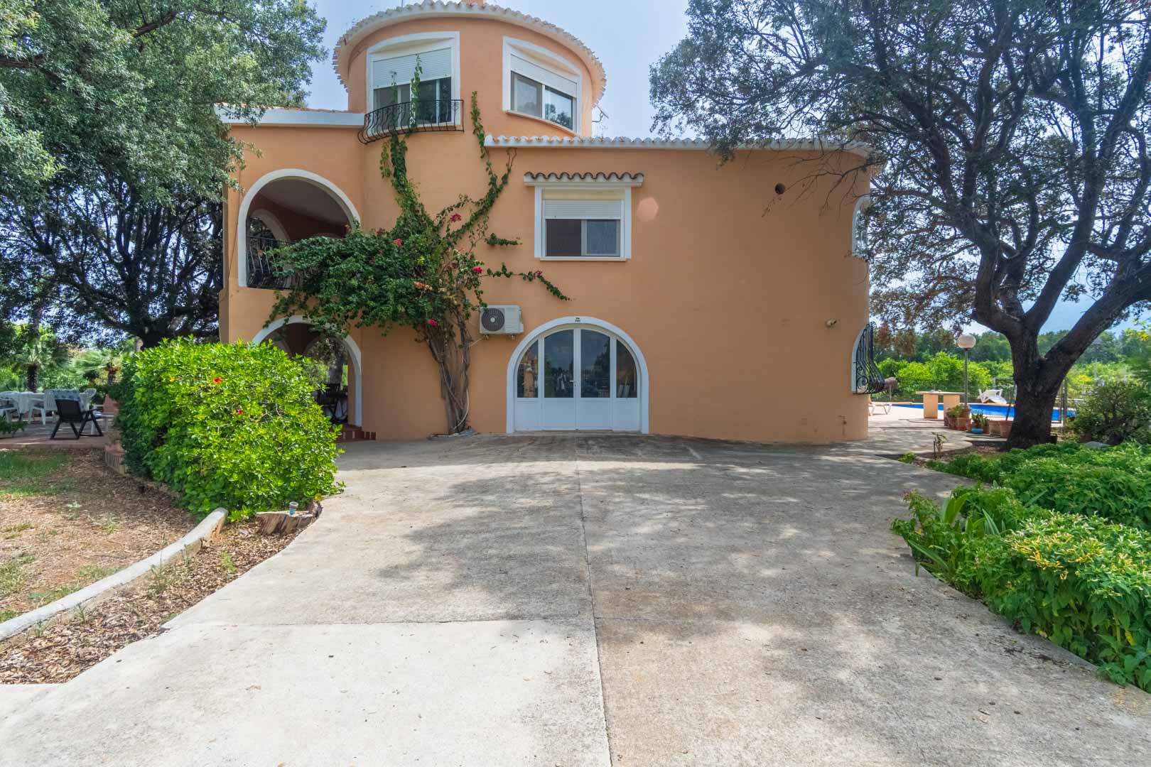Three-story Mediterranean facade featuring a circular tower, arched entrances, and a concrete driveway surrounded by mature trees and climbing plants.