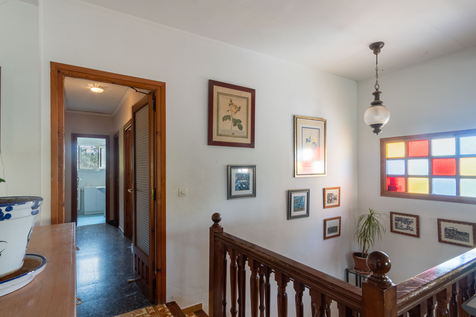 Upper floor landing featuring a turned wood balustrade, colorful stained-glass window, and hallway leading to bedrooms with natural wood door frames.