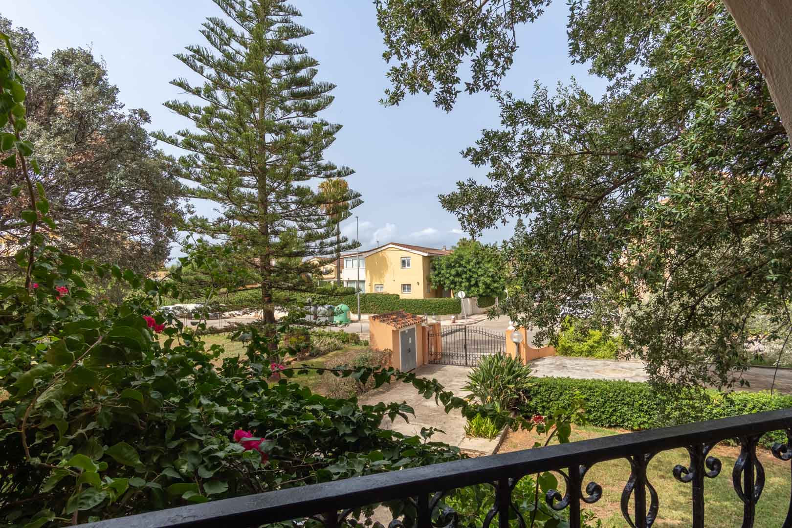 View from the upper terrace featuring a wrought iron railing, overlooking the private garden with mature pines and bougainvillea towards the gated entrance.