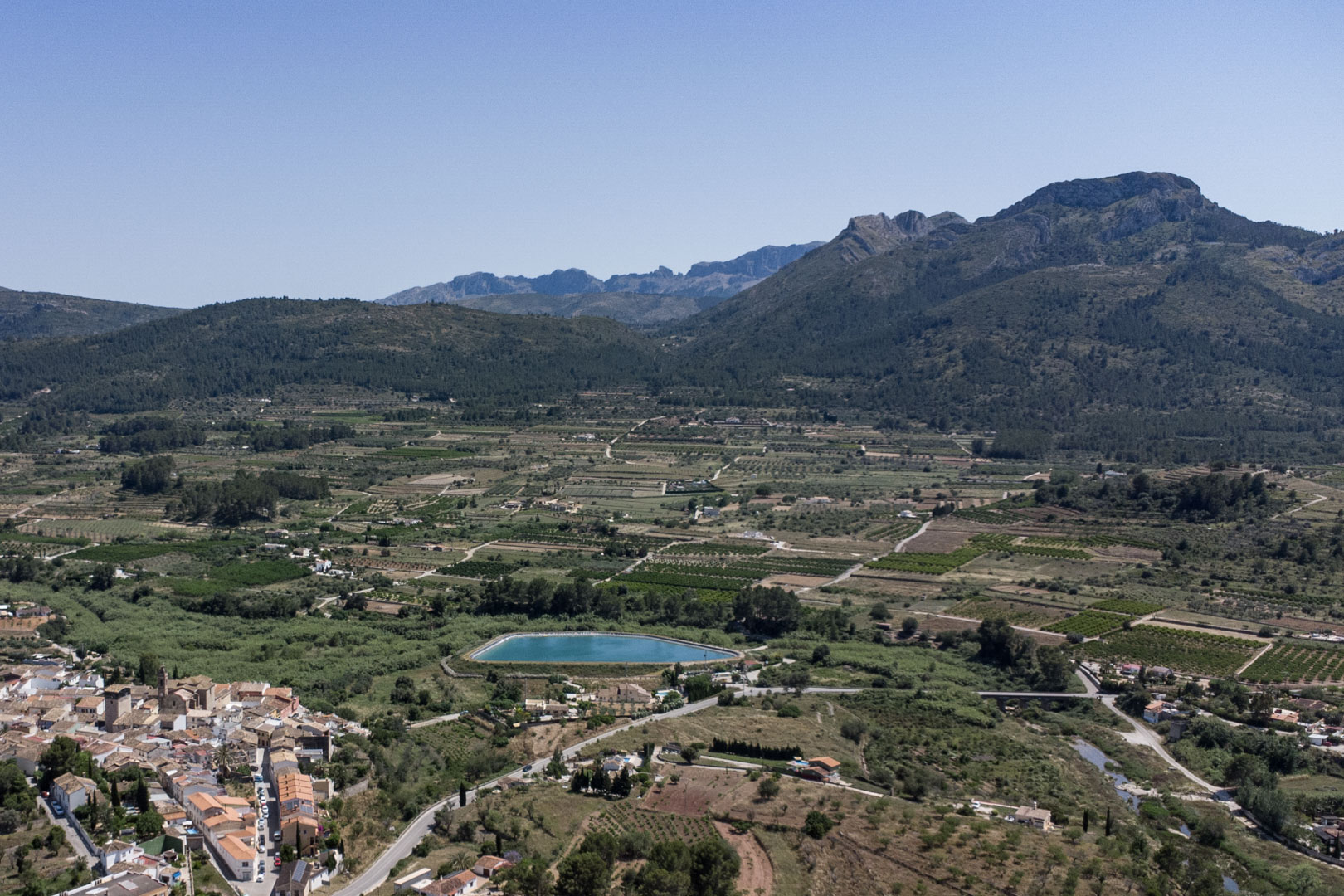 Panoramic aerial view of the Jalón Valley from Alcalalí, featuring agricultural landscapes, a turquoise water reservoir, and the surrounding mountain ranges.
