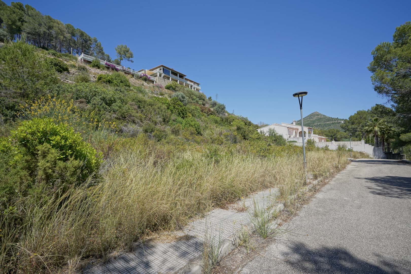 View of an elevated building plot in Alcalalí, showing paved access roads, sidewalks, and street lighting amidst natural hillside vegetation and existing villas.