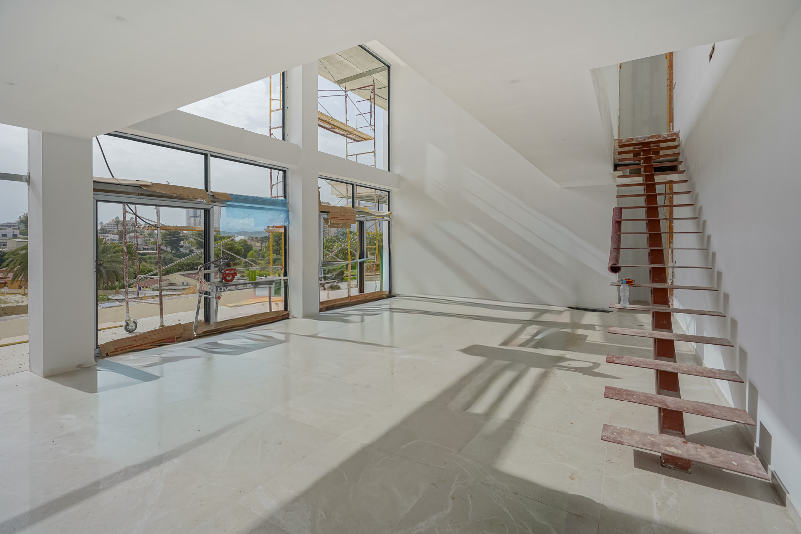 Double-height living area under construction featuring floor-to-ceiling windows, polished stone flooring, and a temporary metal staircase structure.