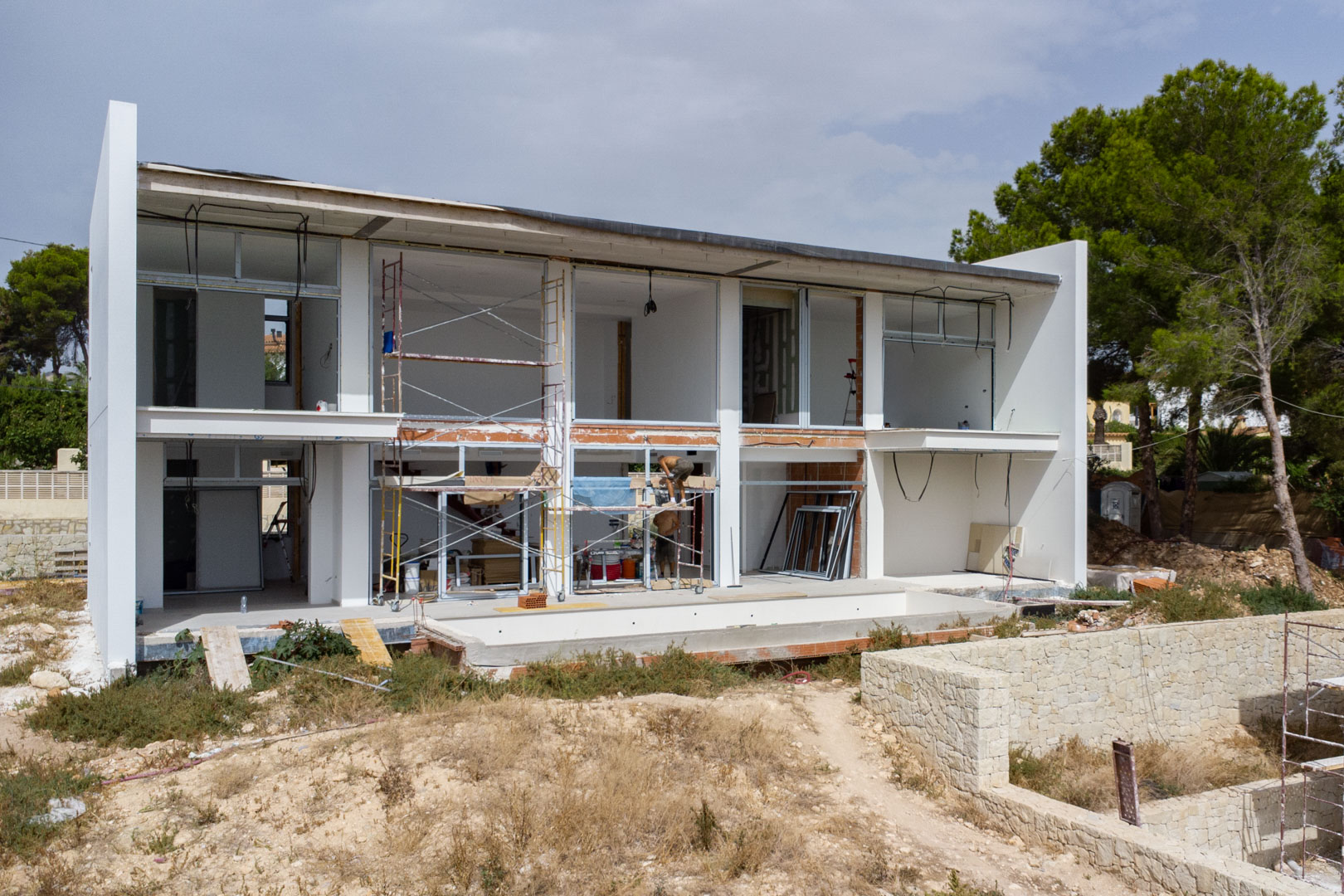 Exterior view of the villa under construction, showing the two-story white facade, large window openings, and the pool basin area.