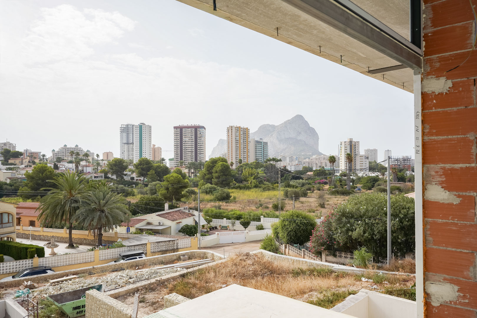 Panoramic view from the upper floor under construction, overlooking the Peñón de Ifach and Calpe skyline, framed by brickwork and concrete slabs.