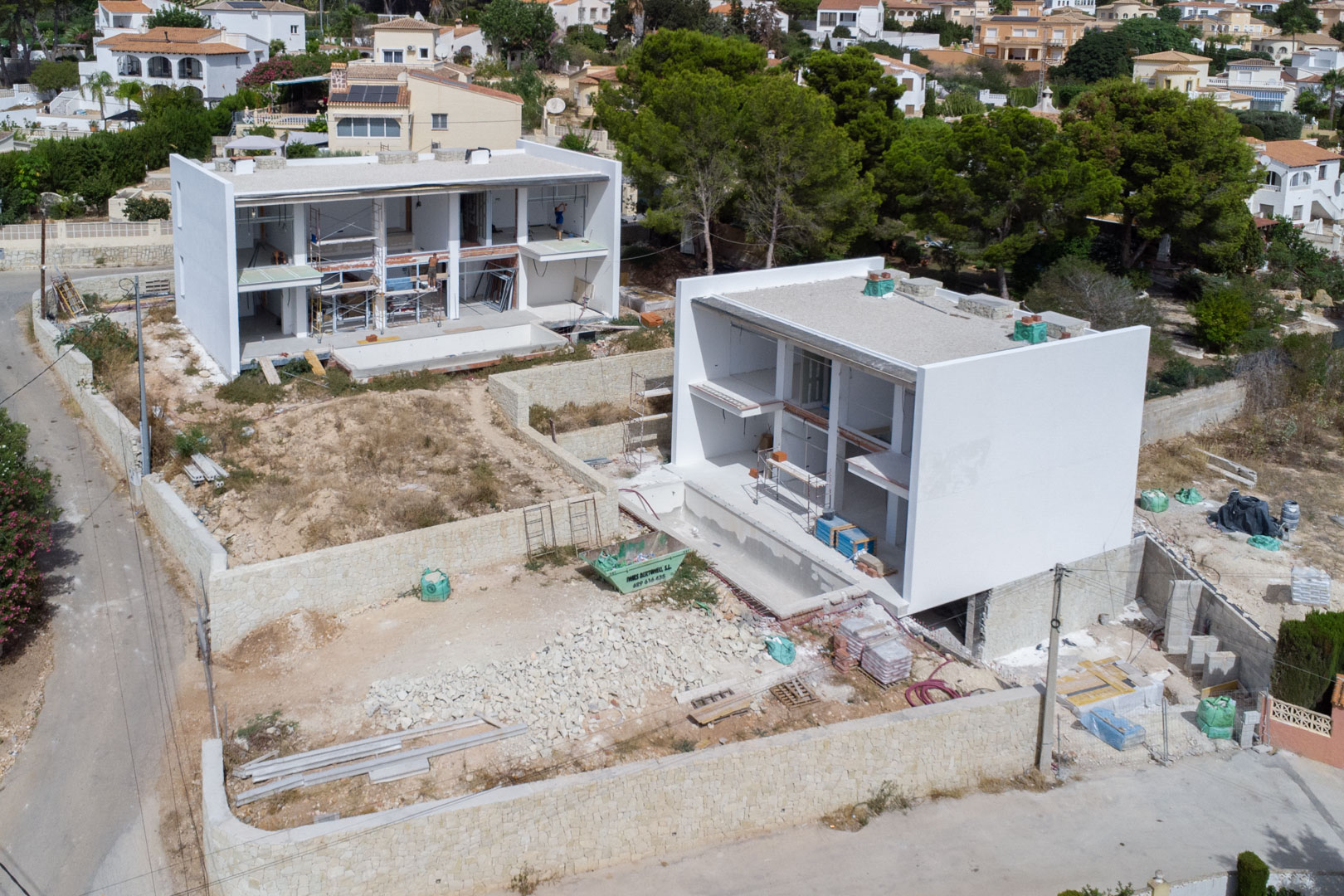 Aerial view showing the current construction progress of the two modern villas, featuring white structural frames, natural stone boundary walls, and site preparation.
