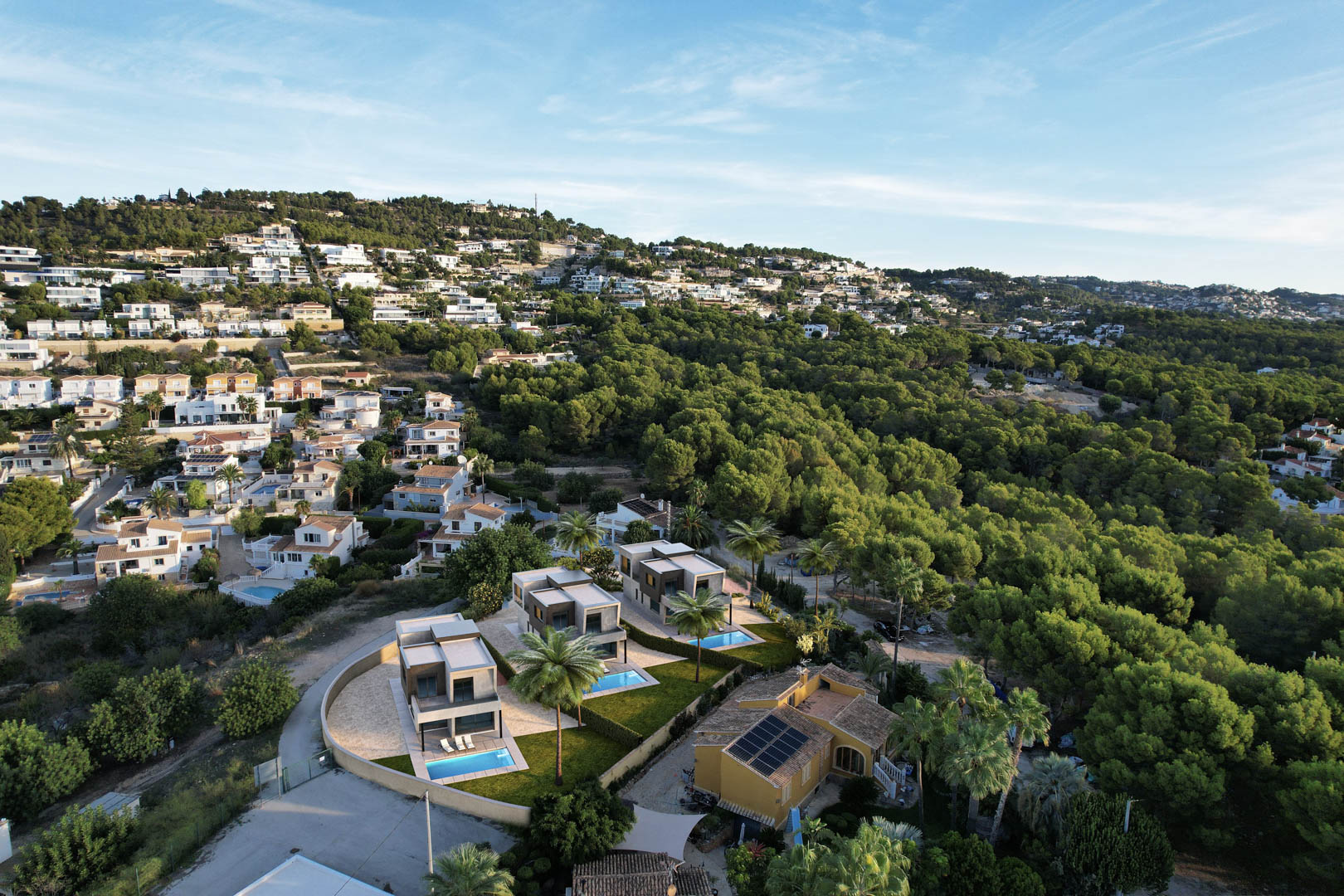 Aerial view of three modern villas featuring private pools and lawns, situated on a hillside adjacent to a lush pine forest in Calpe.