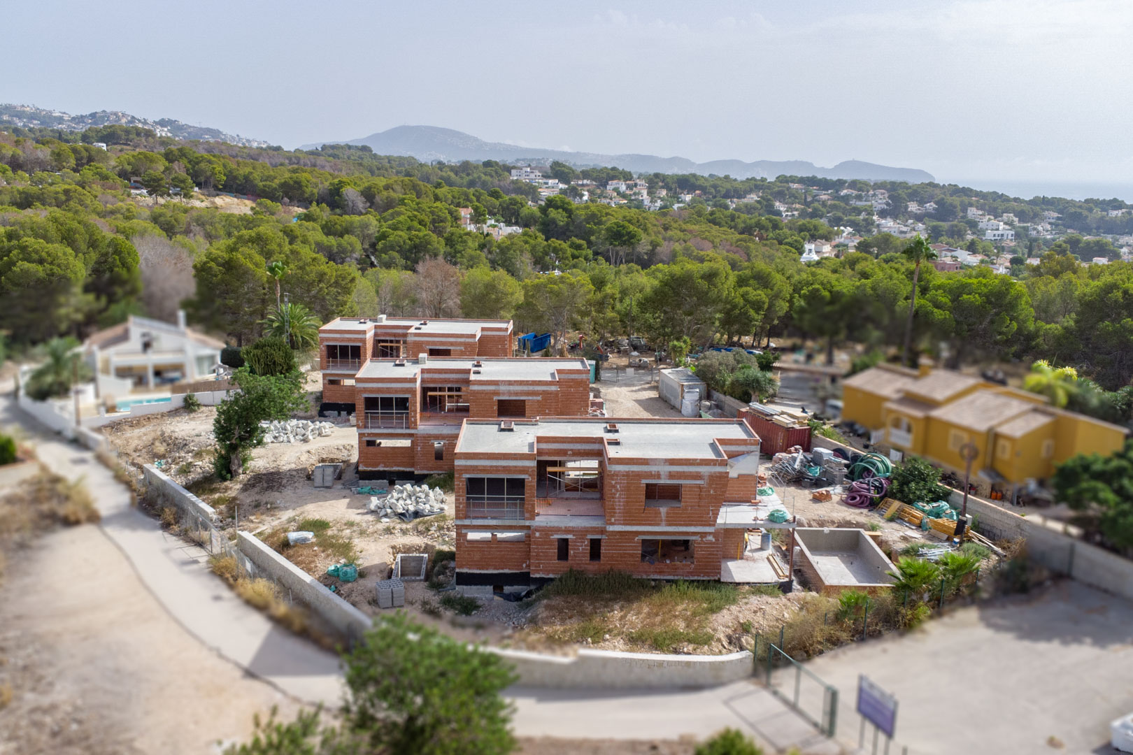 Aerial view showing the construction progress of the three villas, featuring the brick structures, flat roofs, and surrounding Mediterranean pine forest.