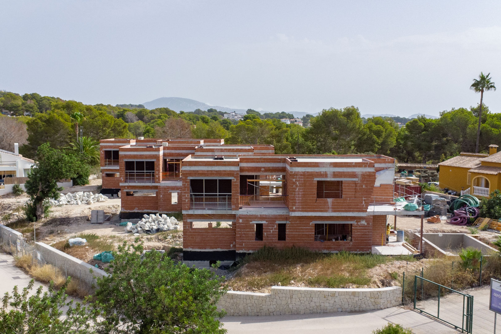 Exterior view of the construction progress, featuring the two-story red brick structures and natural stone perimeter walls in a green setting.