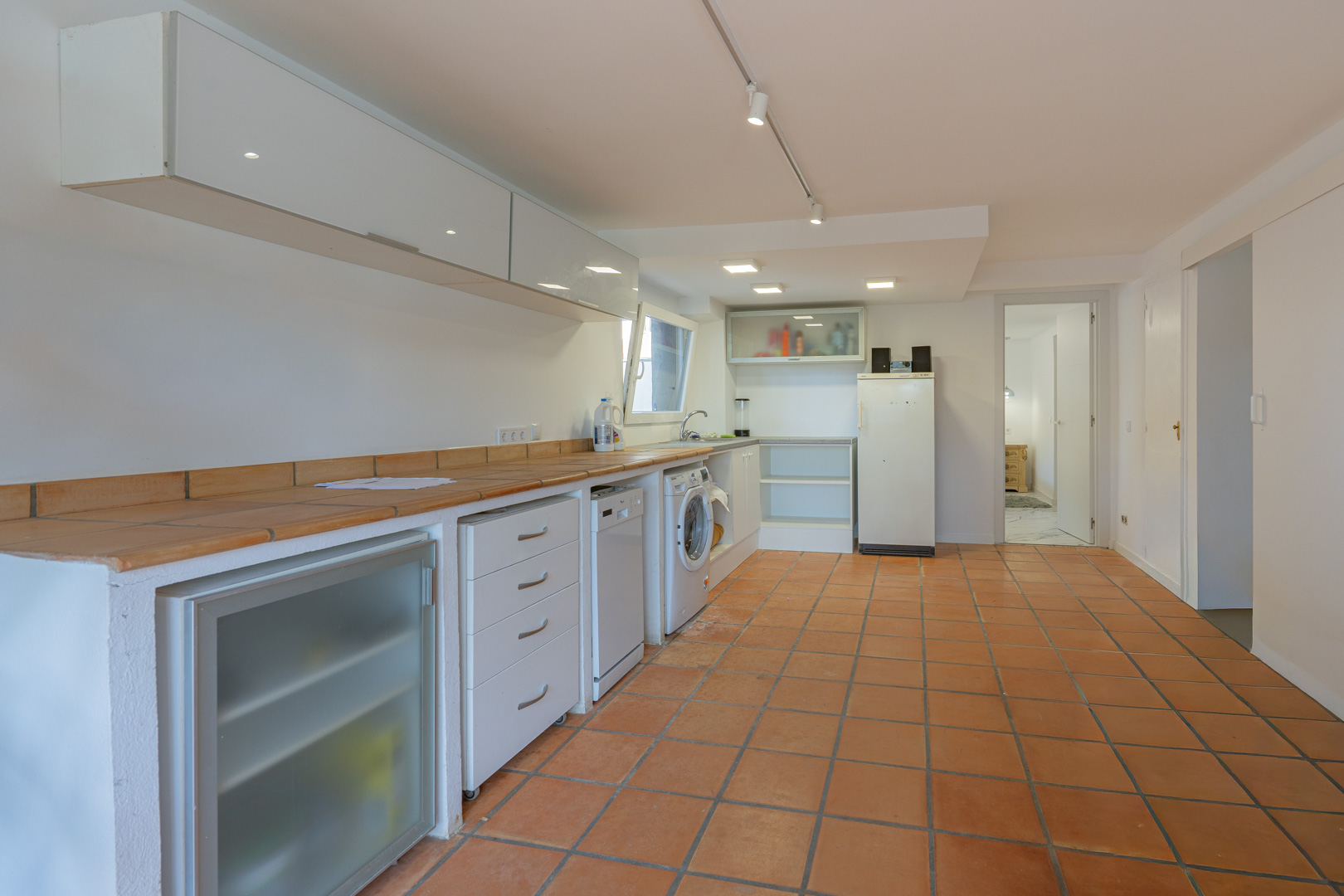 Additional kitchen and laundry area featuring terracotta tile flooring, tiled countertops, and integrated appliances beneath minimalist white cabinetry.