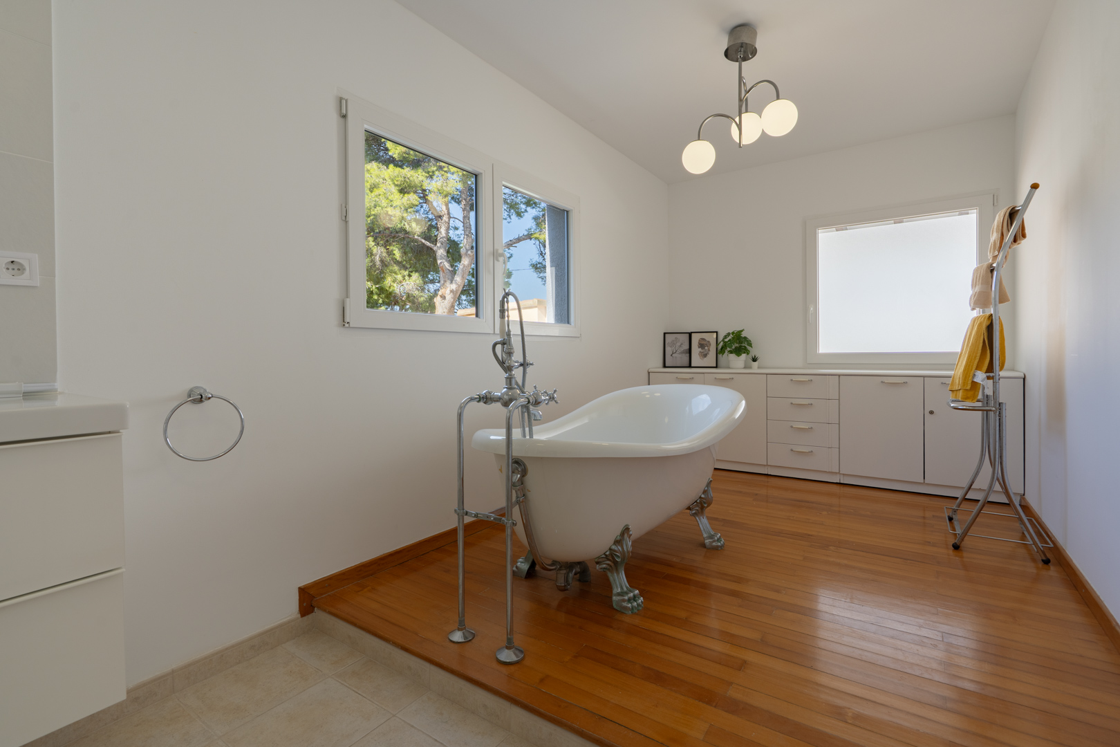 Bathroom featuring a freestanding clawfoot bathtub on a raised wooden floor, complemented by white cabinetry and natural light from large windows.