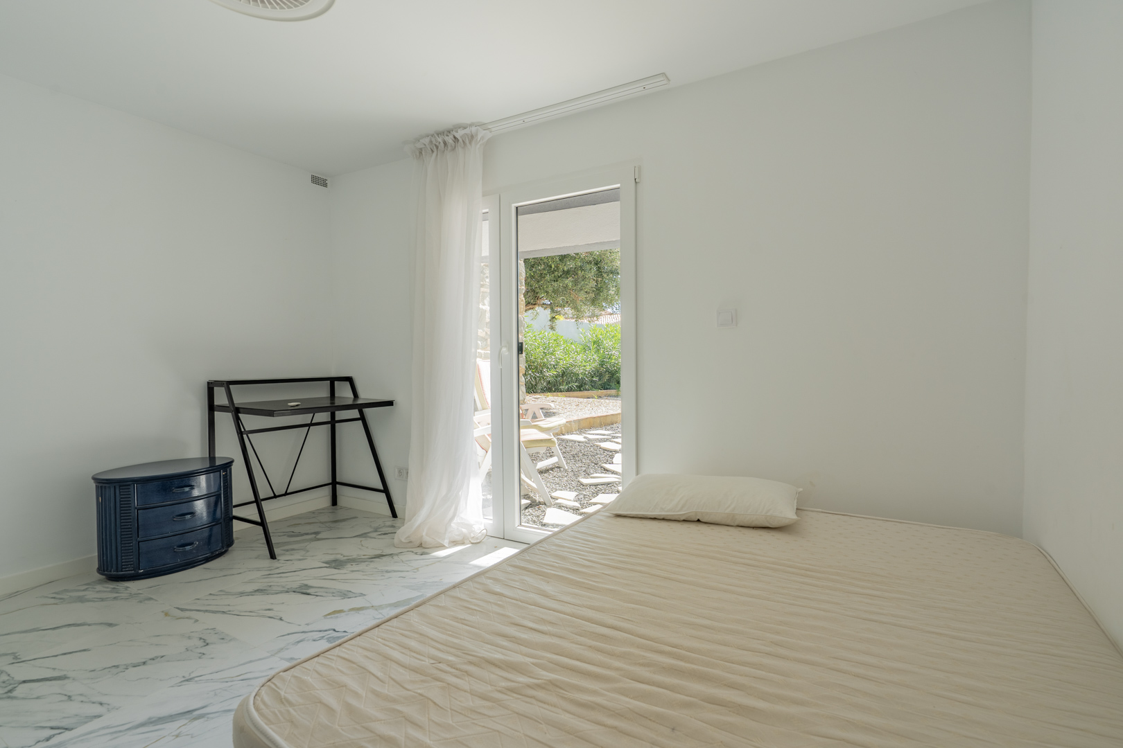 Bedroom featuring marble-patterned flooring and direct access to the garden terrace. Includes a minimalist desk and a blue chest of drawers.