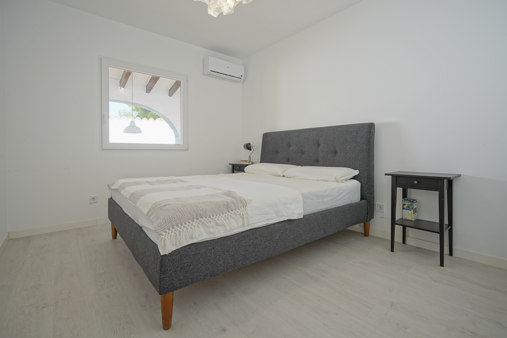 Bright bedroom featuring a grey upholstered double bed, split-unit air conditioning, and light laminate flooring. The window overlooks a covered terrace with traditional beams.