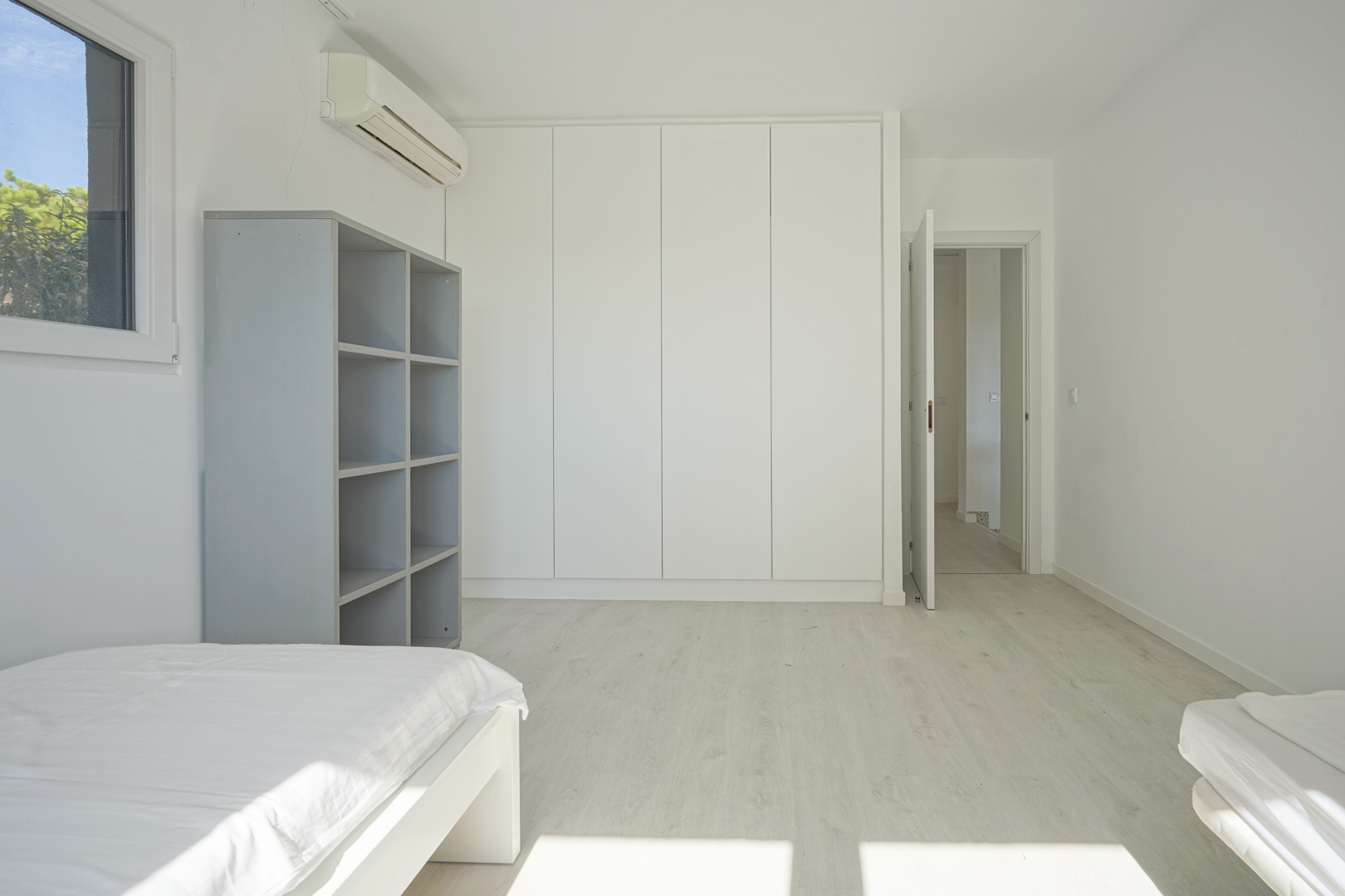 Bright bedroom featuring floor-to-ceiling white built-in wardrobes, light laminate flooring, wall-mounted air conditioning, and a grey open shelving unit.