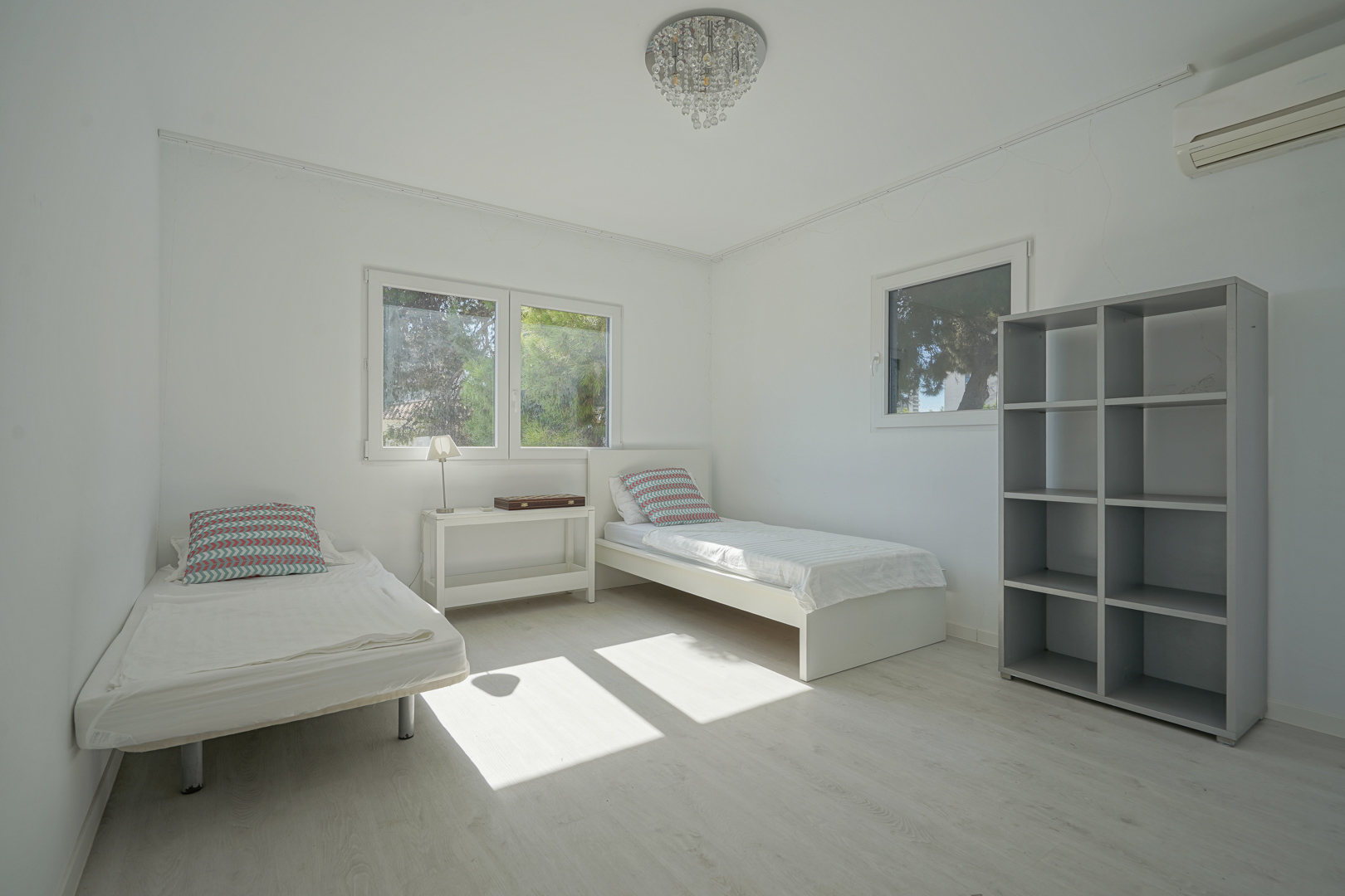 Bright bedroom featuring two single beds, light wood flooring, and a grey shelving unit. Includes wall-mounted AC, a crystal chandelier, and windows overlooking greenery.
