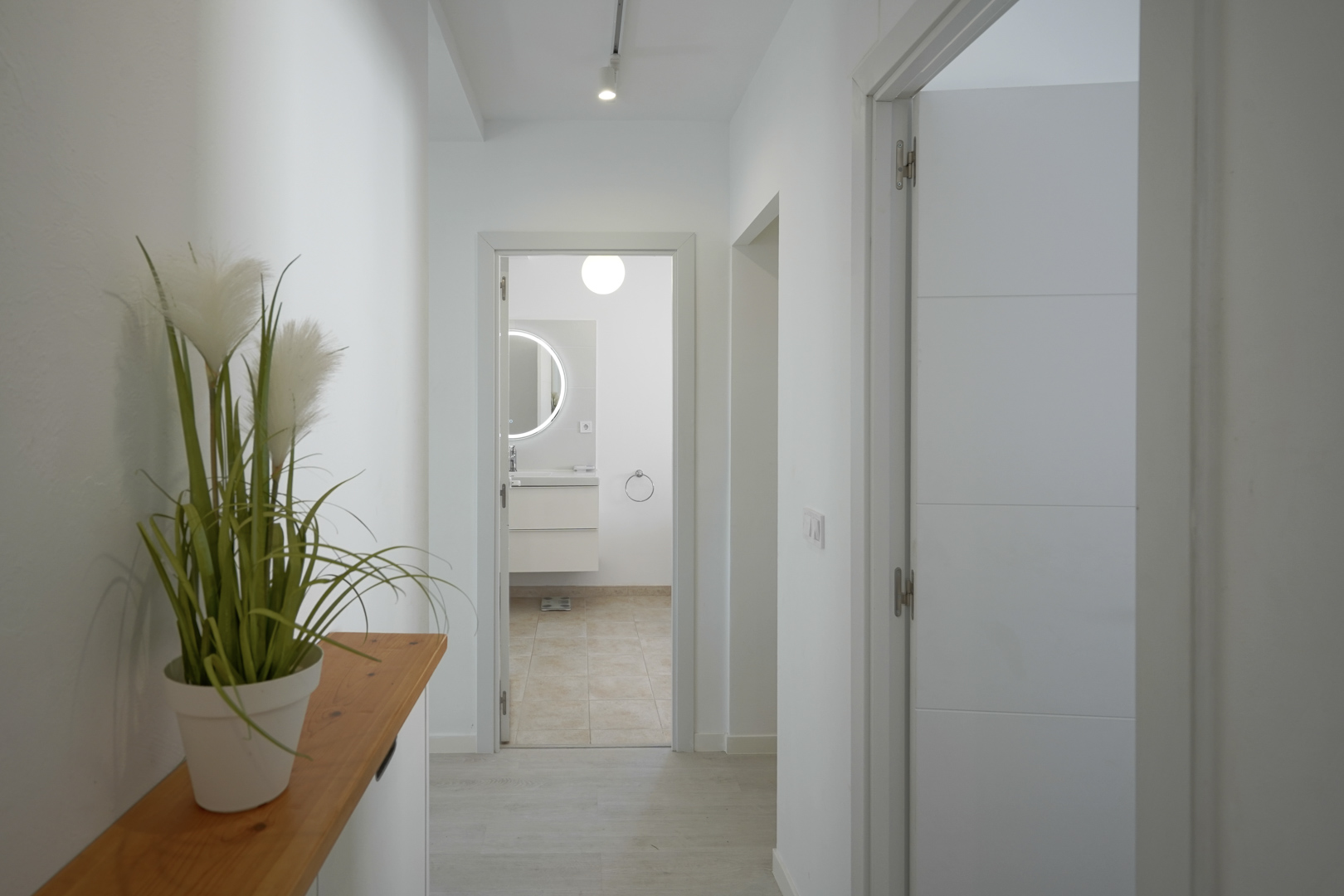 Bright hallway with white walls and light flooring leading to a modern bathroom featuring a backlit circular mirror and floating vanity.