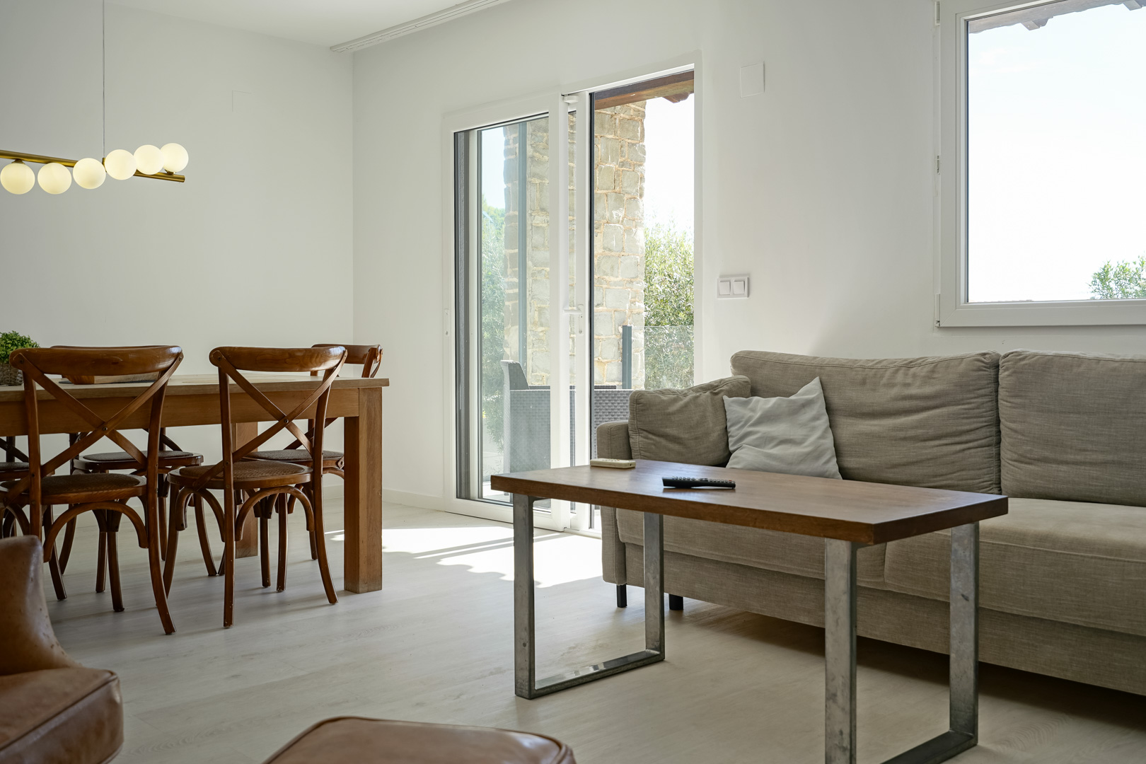 Bright living-dining area with wooden furniture and a globe light fixture. Double-glazed doors connect the interior to the natural stone terrace.