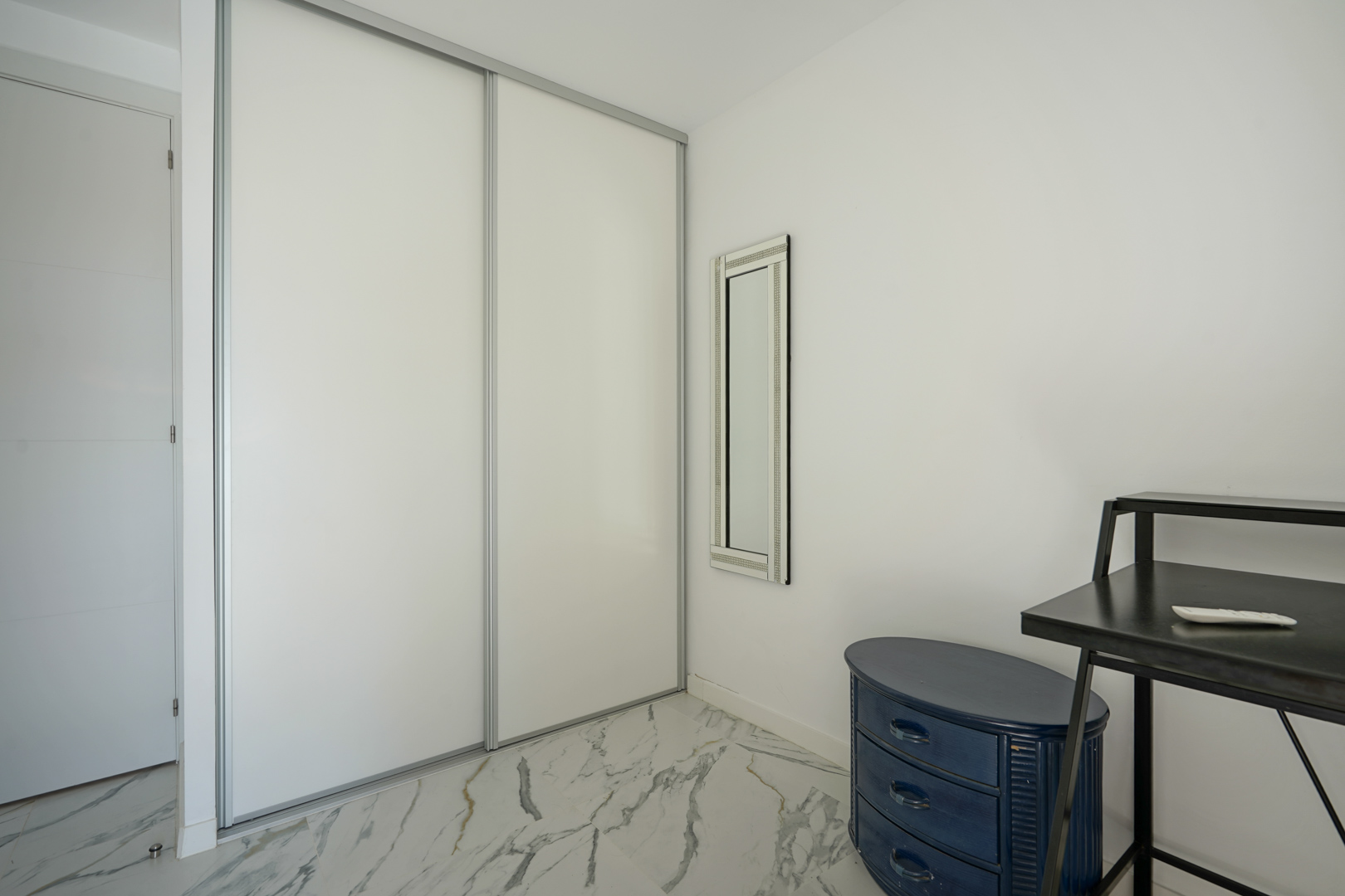 Built-in wardrobe with white sliding doors set against marble flooring, featuring a vertical mirror and a blue oval chest of drawers.