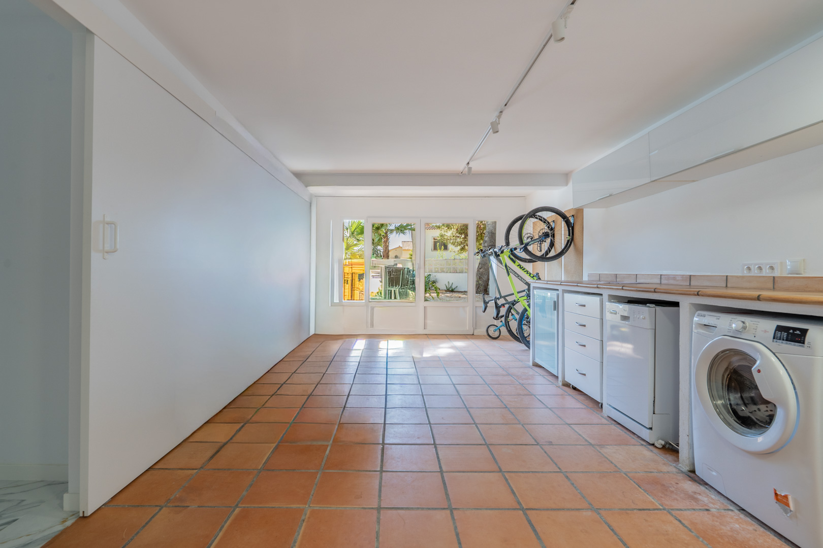 Converted garage featuring terracotta tile flooring, a functional laundry area with integrated appliances, storage cabinets, and a glass door to the garden.
