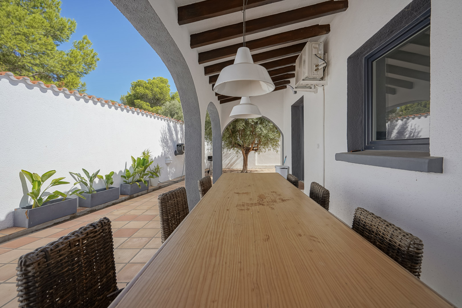 Covered terrace featuring a long wooden dining table, wicker chairs, and exposed ceiling beams. The arched architecture overlooks a Mediterranean garden with an olive tree.