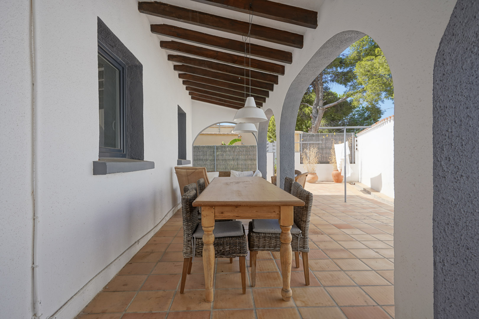 Covered terrace featuring exposed wooden beams, Mediterranean arches, and an outdoor dining area with a wooden table and wicker chairs on terracotta tiles.