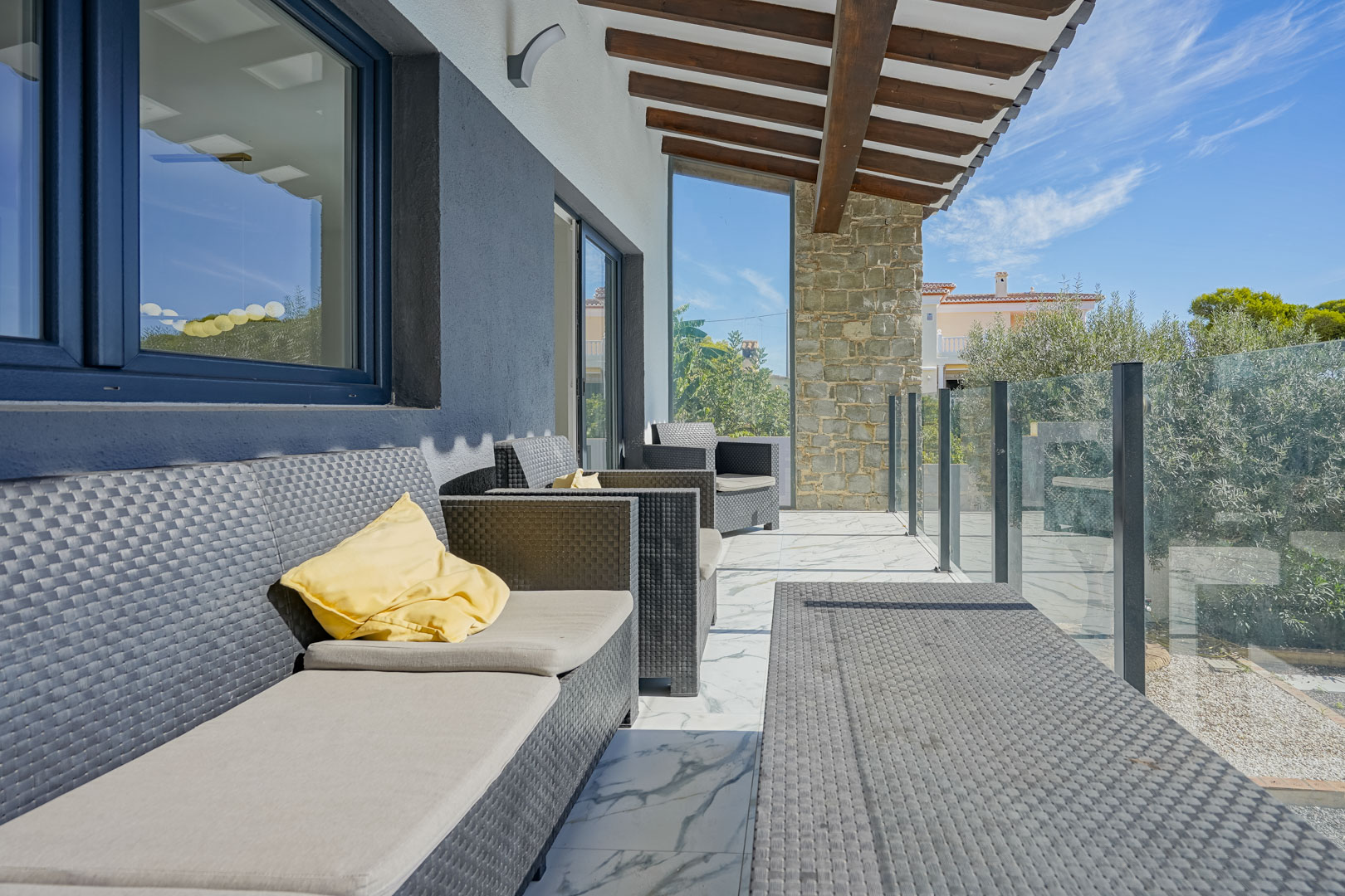 Covered terrace featuring exposed wooden beams, synthetic rattan lounge furniture, and marble-effect flooring. A glass balustrade offers views of the surrounding greenery.