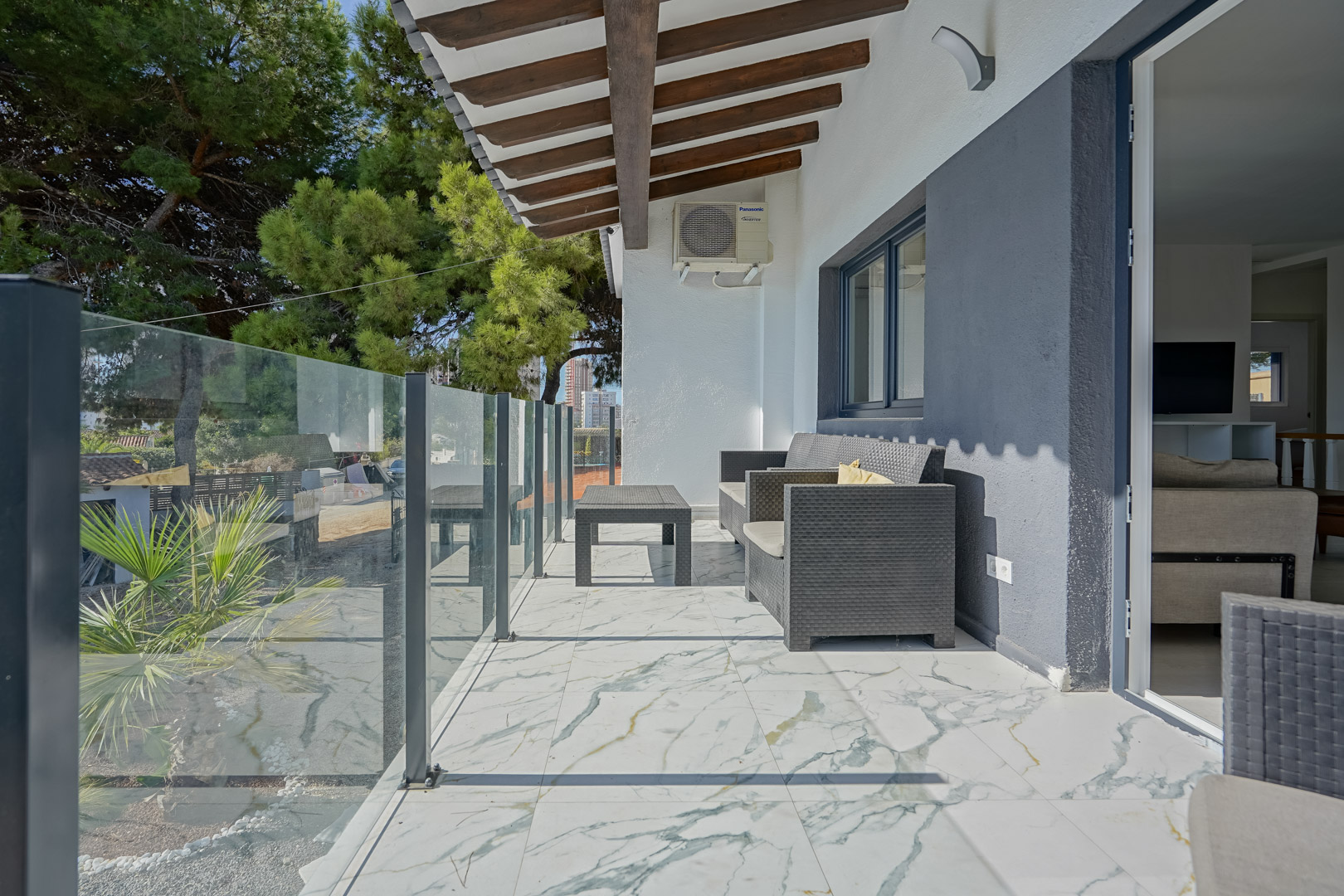 Covered terrace featuring marble-effect tiling, a glass balustrade, and a wooden beam ceiling. Equipped with grey rattan seating and views of the Mediterranean greenery.