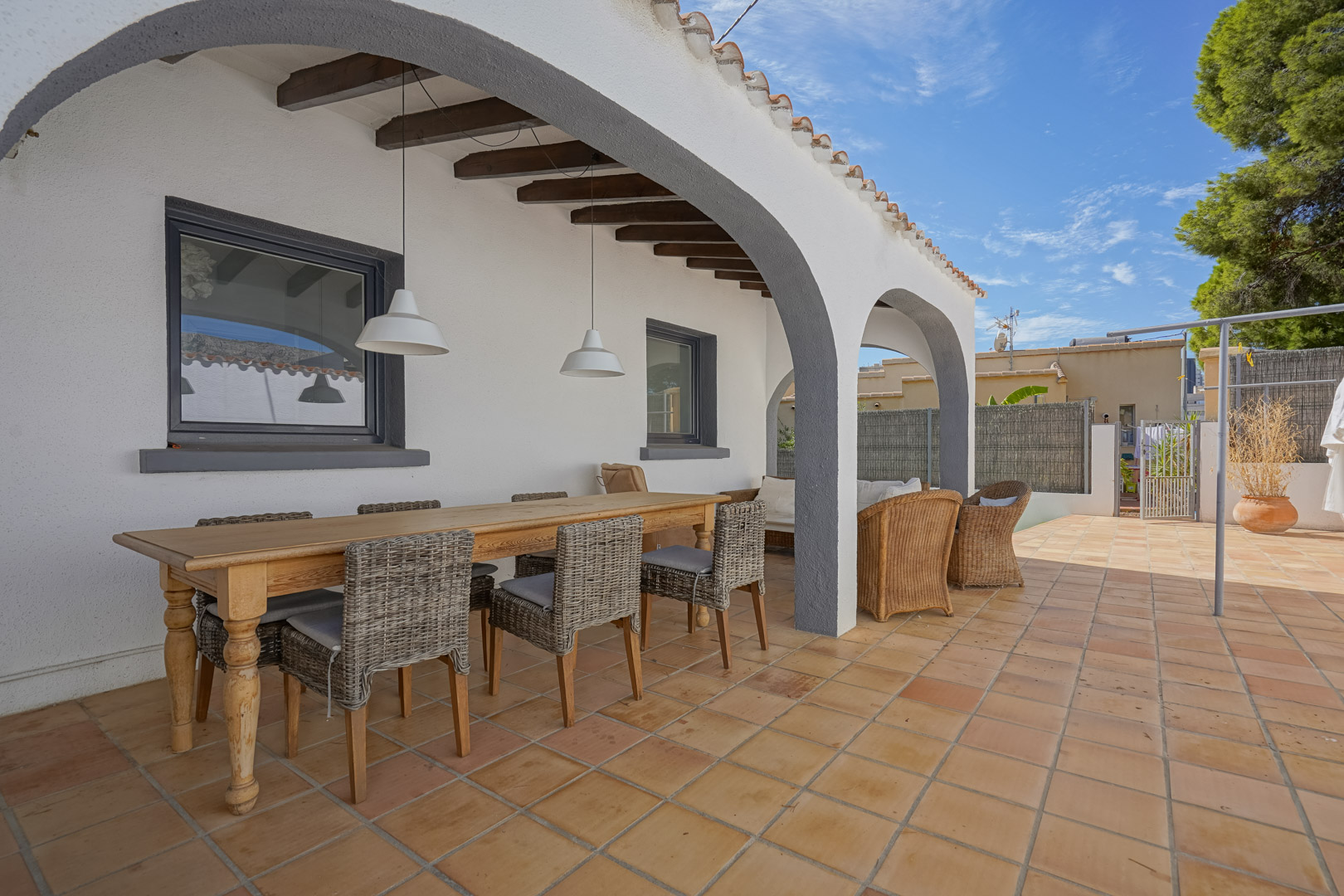 Covered terrace featuring white arches, exposed wooden beams, and terracotta tiling. It includes a dining area with a wooden table and wicker chairs.