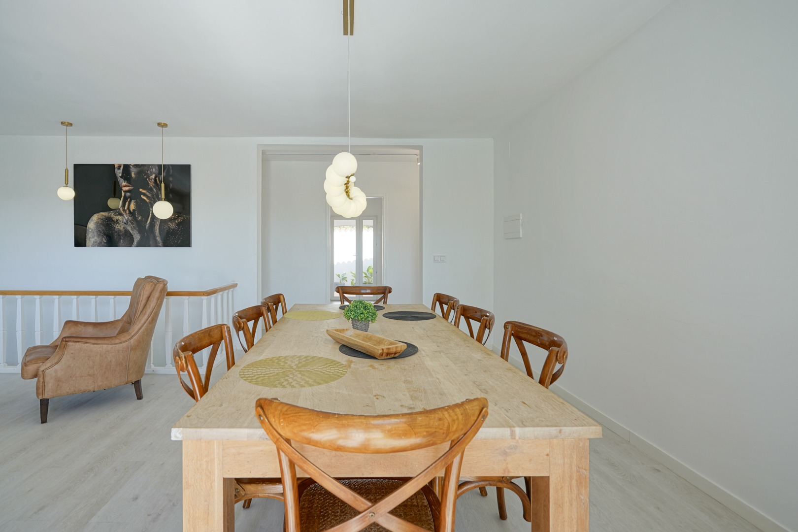 Dining area featuring a large light wood table with X-back chairs, modern globe pendant lighting, and a leather armchair adjacent to a decorative wall painting in an open layout.