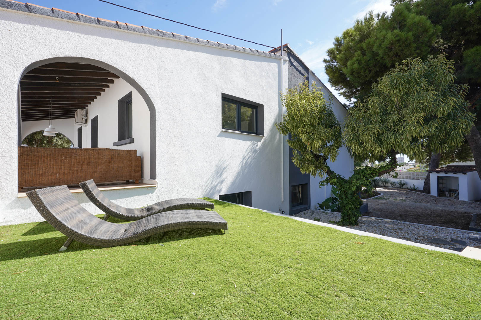Elevated lawn area featuring curved wicker sun loungers adjacent to the white facade and a covered terrace with dark wooden beams.