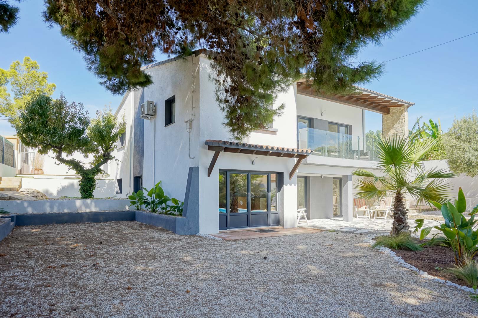 Exterior facade of the villa featuring grey window frames, glass-railed terraces, and a Mediterranean garden with gravel and mature pine trees.