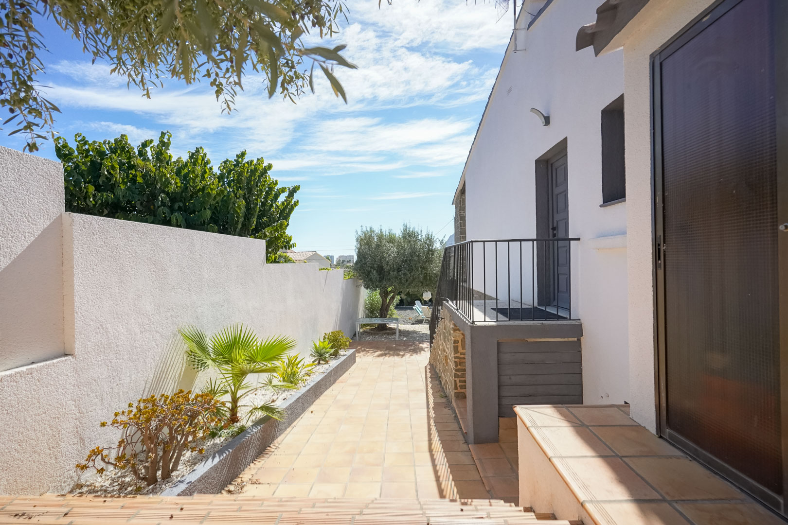 Exterior tiled walkway featuring a white perimeter wall with Mediterranean plantings, leading to a side entrance with stone accents and a black metal railing.