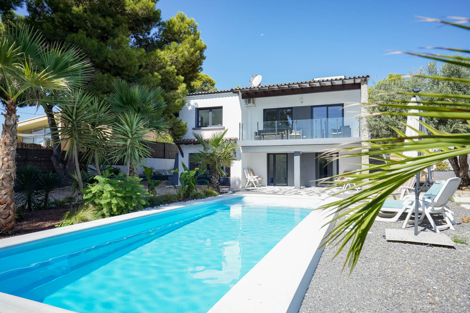 Exterior view featuring a rectangular swimming pool, Mediterranean landscaping with palms, and a two-story white villa with glass-railed balconies.